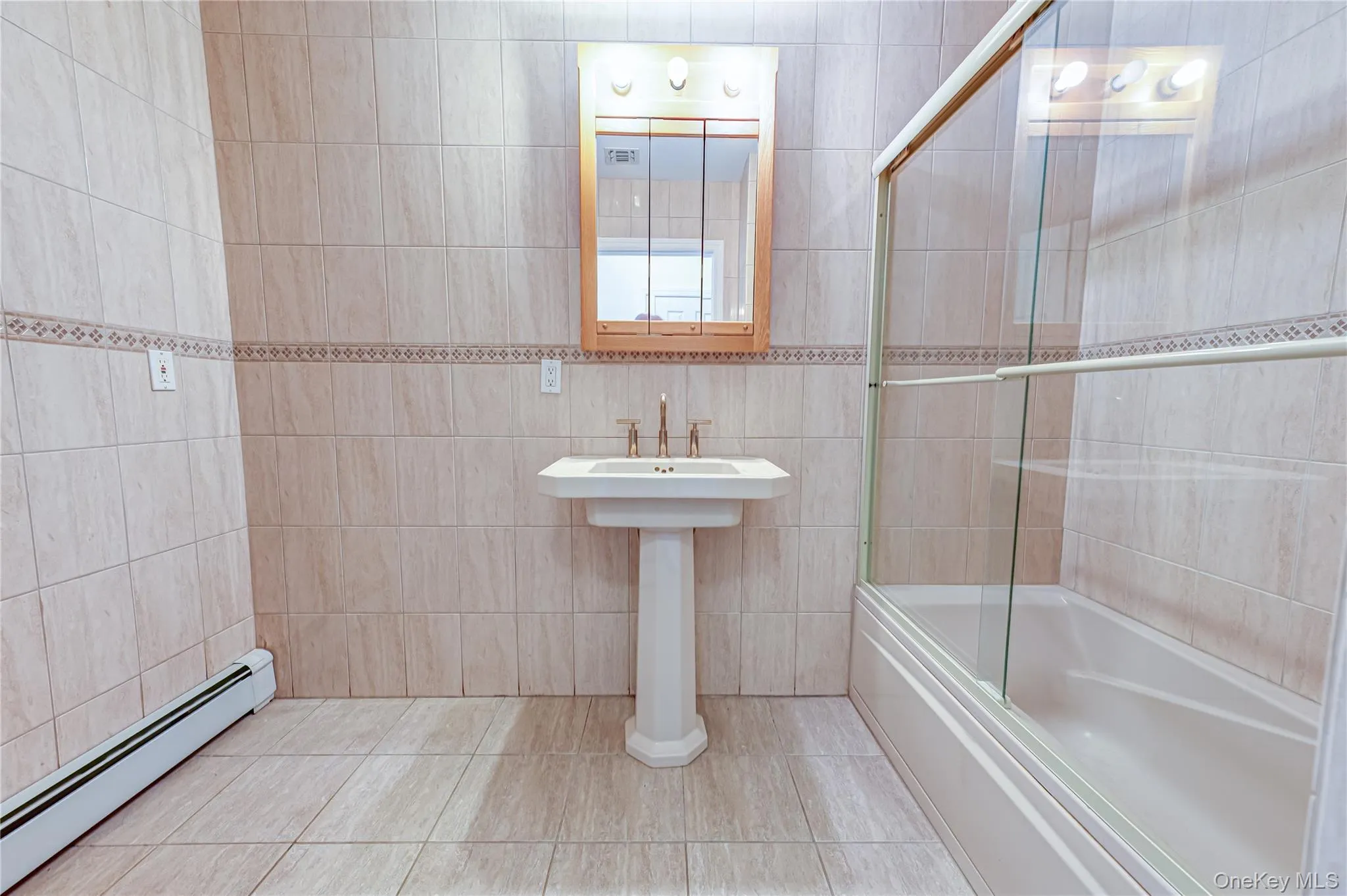 Pedestal sink with chrome faucet, full tile flooring, tiled walls with decorative band, bathtub with glass sliding enclosure, and wood-framed medicine cabinet Pedestal sink with chrome faucet, full tile flooring, tiled walls with decorative band, bathtub with glass sliding enclosure, and wood-framed medicine cabinet