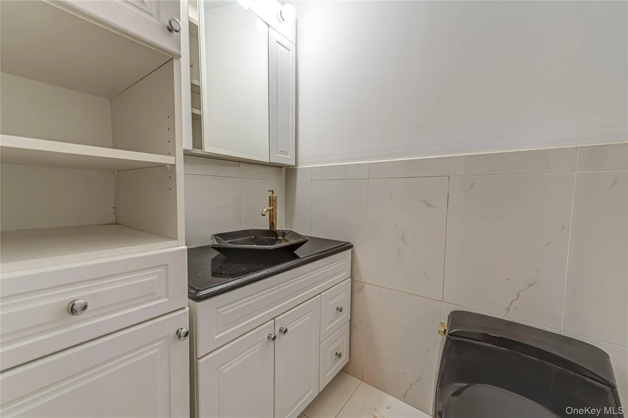 1/2 bathroom, White shaker cabinetry with brushed nickel hardware, black countertop, modern vessel sink with a gold-finish faucet, white tiled walls with subtle veining, and a black toilet 1/2 bathroom, White shaker cabinetry with brushed nickel hardware, black countertop, modern vessel sink with a gold-finish faucet, white tiled walls with subtle veining, and a black toilet