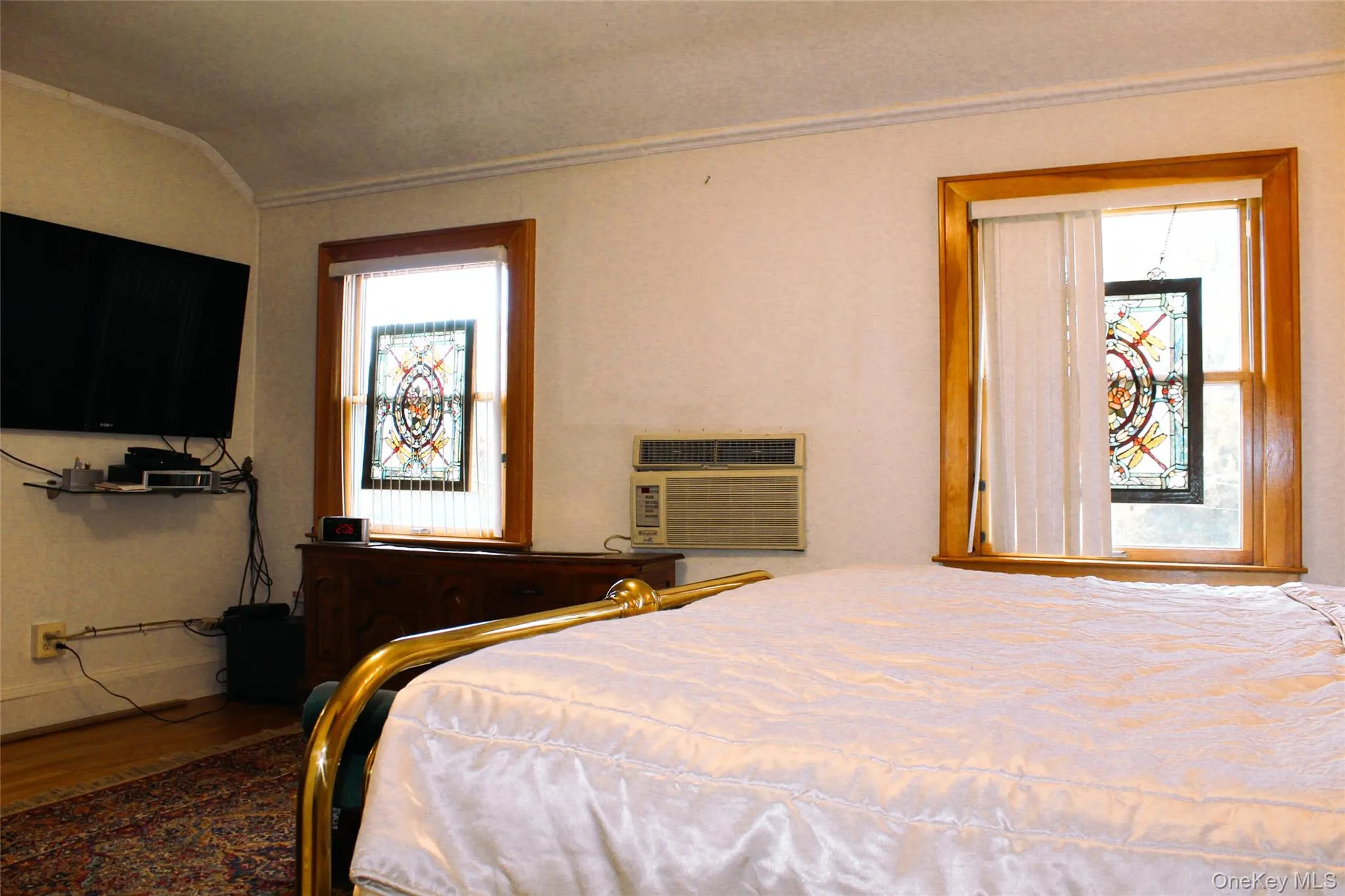 Large bedroom 1 with wood-frame windows with stained glass decorative inserts, white textured walls, and a wall-mounted air conditioning unit Large bedroom 1 with wood-frame windows with stained glass decorative inserts, white textured walls, and a wall-mounted air conditioning unit