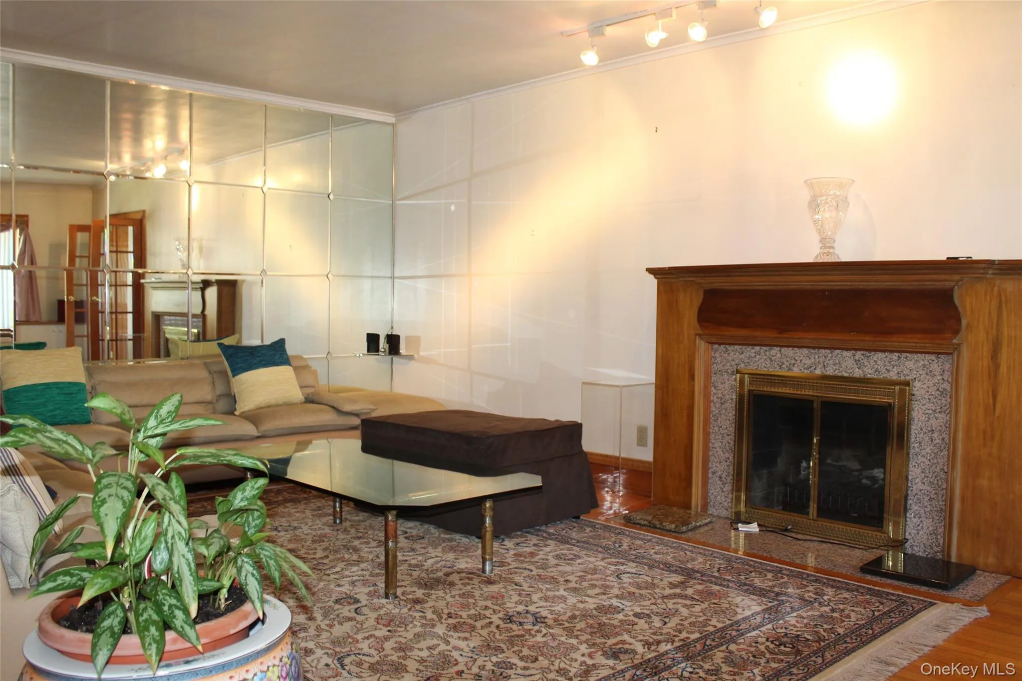 Living room has wood-finish flooring with a working fireplace featuring a wood mantel and stone surround, and a wall of mirrored panels Living room has wood-finish flooring with a working fireplace featuring a wood mantel and stone surround, and a wall of mirrored panels