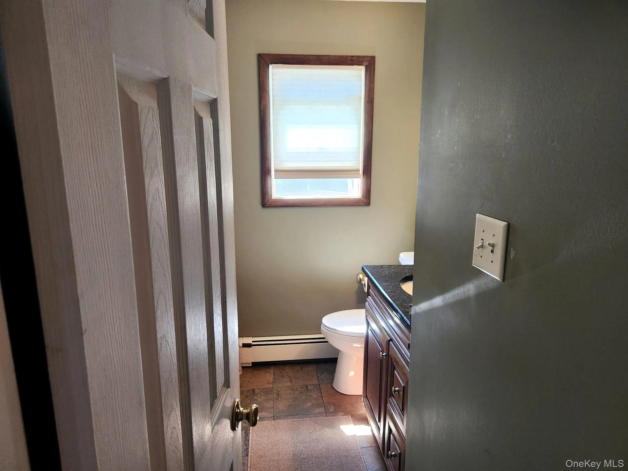 Bathroom featuring a dark wood finish vanity with a dark countertop, white toilet, and a window with a blind Bathroom featuring a dark wood finish vanity with a dark countertop, white toilet, and a window with a blind
