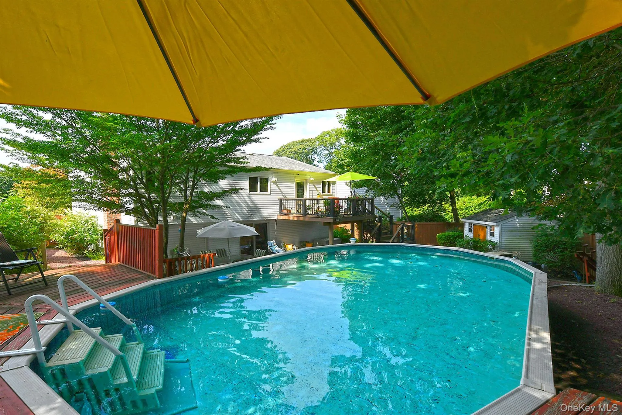 Expansive pool area featuring a curved pool with integrated steps, a surrounding wood deck, mature trees, and a detached storage shed Expansive pool area featuring a curved pool with integrated steps, a surrounding wood deck, mature trees, and a detached storage shed