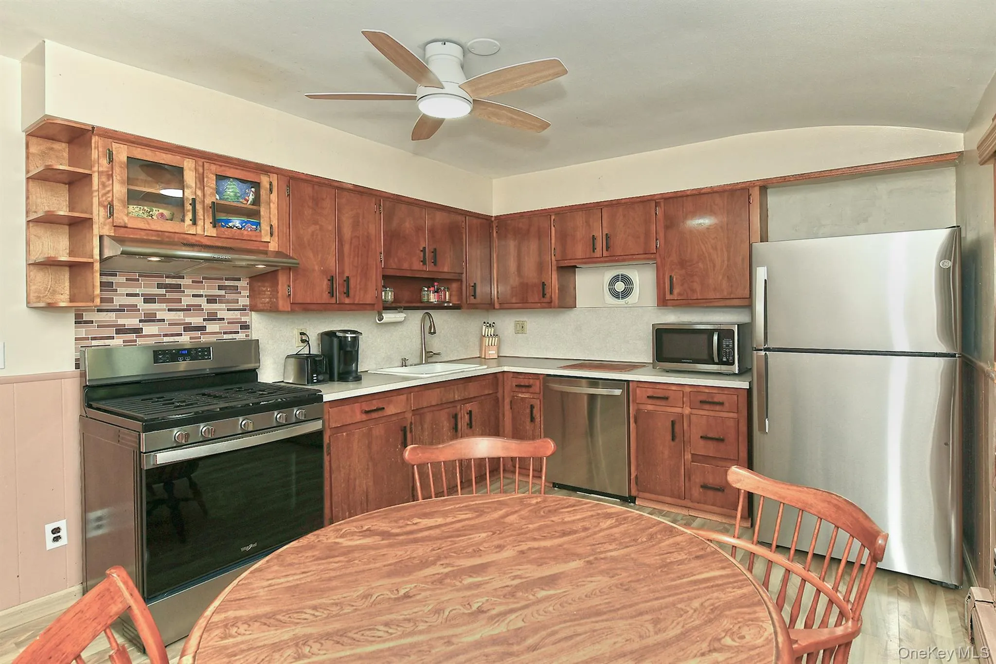 Wood-finish cabinetry, stainless steel appliances, mosaic tile backsplash, solid surface countertops, and a ceiling fan with integrated lighting Wood-finish cabinetry, stainless steel appliances, mosaic tile backsplash, solid surface countertops, and a ceiling fan with integrated lighting