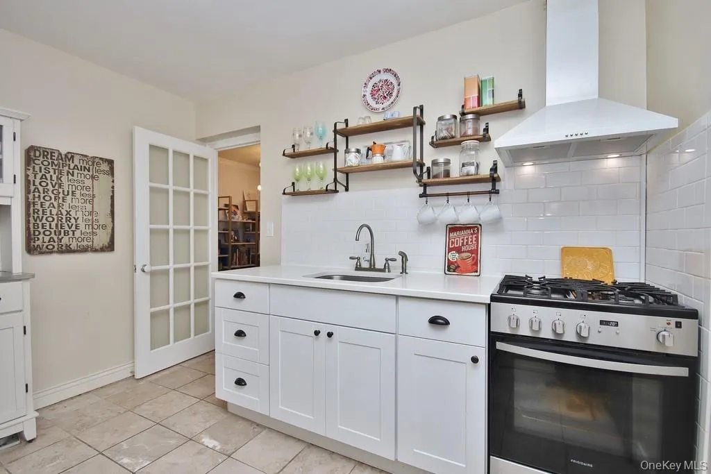 White shaker-style cabinetry with dark hardware, stainless steel range with matching range hood, white subway tile backsplash, light-colored countertop, and a single basin sink with a gooseneck faucet White shaker-style cabinetry with dark hardware, stainless steel range with matching range hood, white subway tile backsplash, light-colored countertop, and a single basin sink with a gooseneck faucet