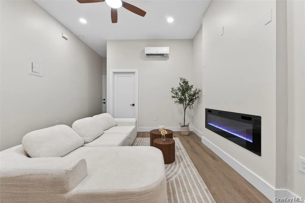Recessed electric fireplace, ceiling fan, wood-finish flooring, white walls, and a mini-split air conditioning unit Recessed electric fireplace, ceiling fan, wood-finish flooring, white walls, and a mini-split air conditioning unit