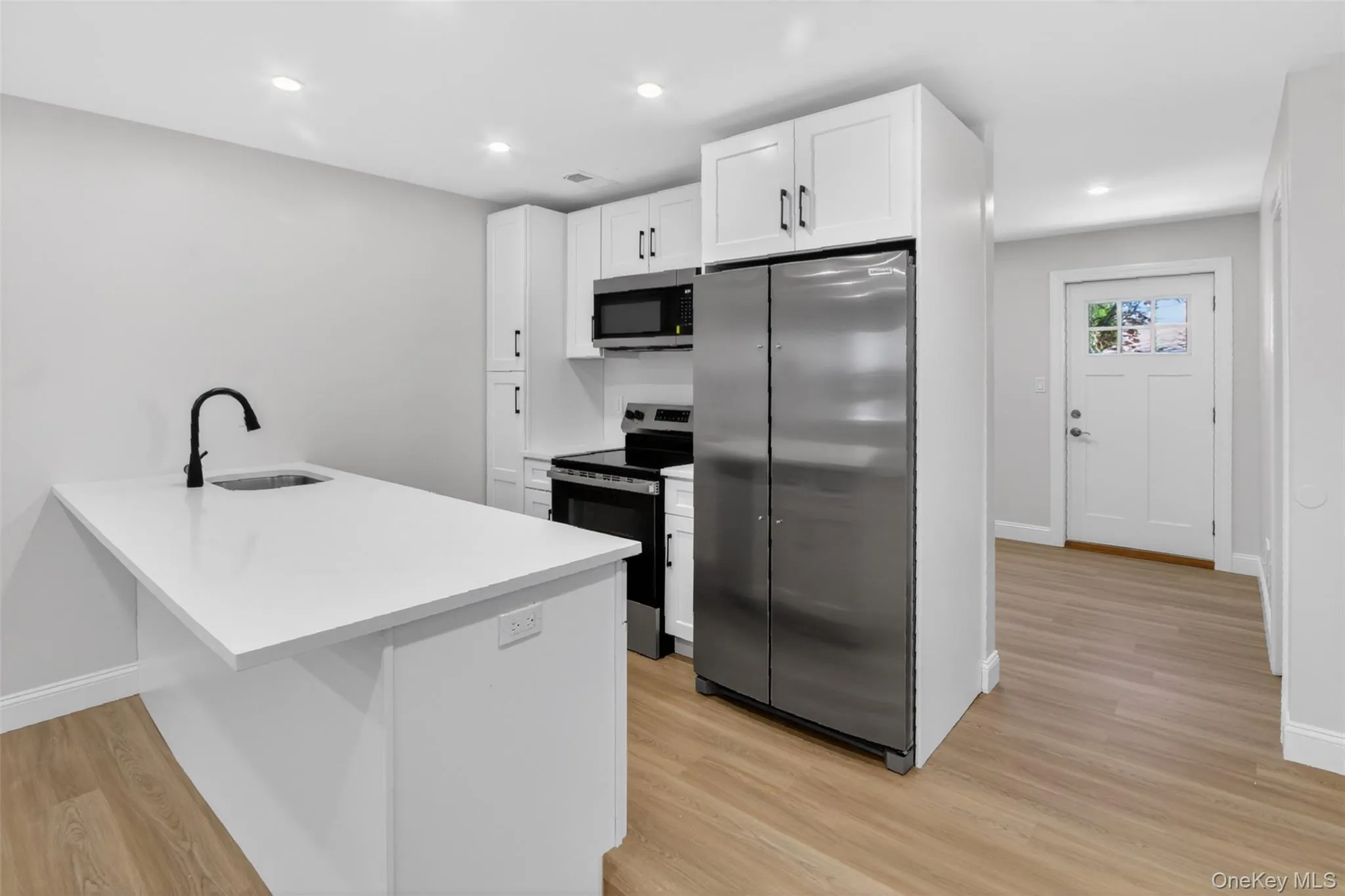 Modern kitchen featuring a large island with sink, white shaker cabinetry, stainless steel appliances, and wood-finish flooring Modern kitchen featuring a large island with sink, white shaker cabinetry, stainless steel appliances, and wood-finish flooring
