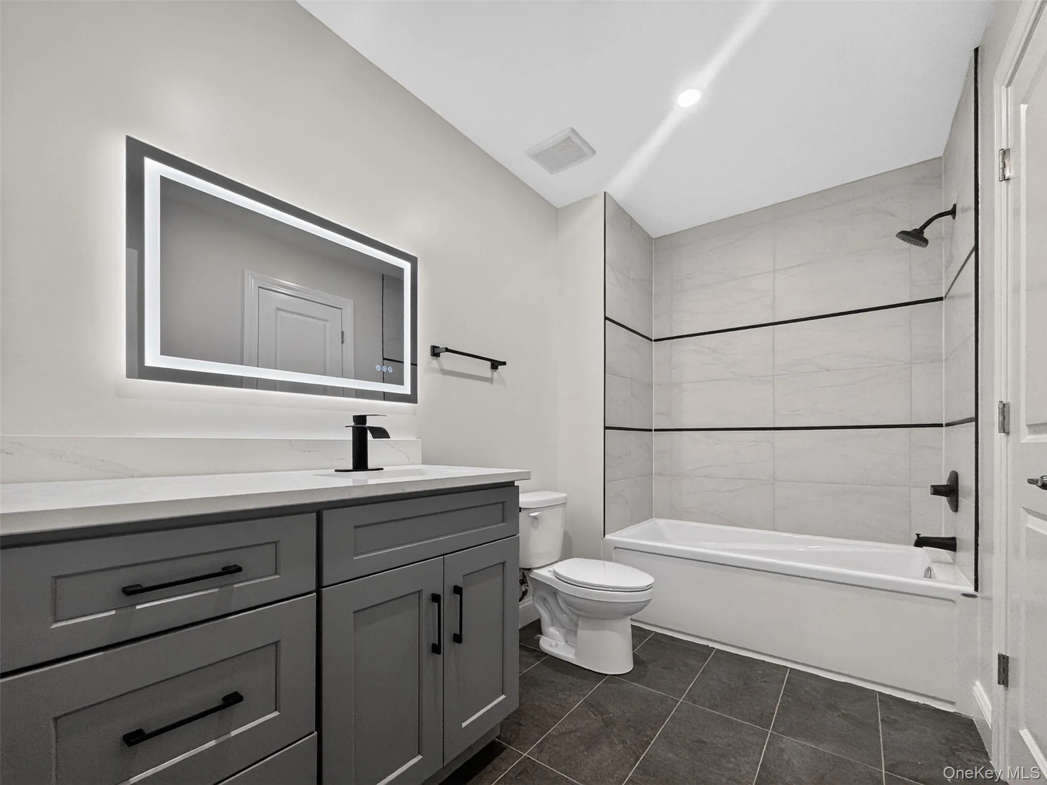 Bathroom featuring a gray vanity with a light-colored countertop, an illuminated rectangular mirror, a white bathtub with light-toned tiled surround and dark accent lines, a white toilet, and dark square floor tiles Bathroom featuring a gray vanity with a light-colored countertop, an illuminated rectangular mirror, a white bathtub with light-toned tiled surround and dark accent lines, a white toilet, and dark square floor tiles
