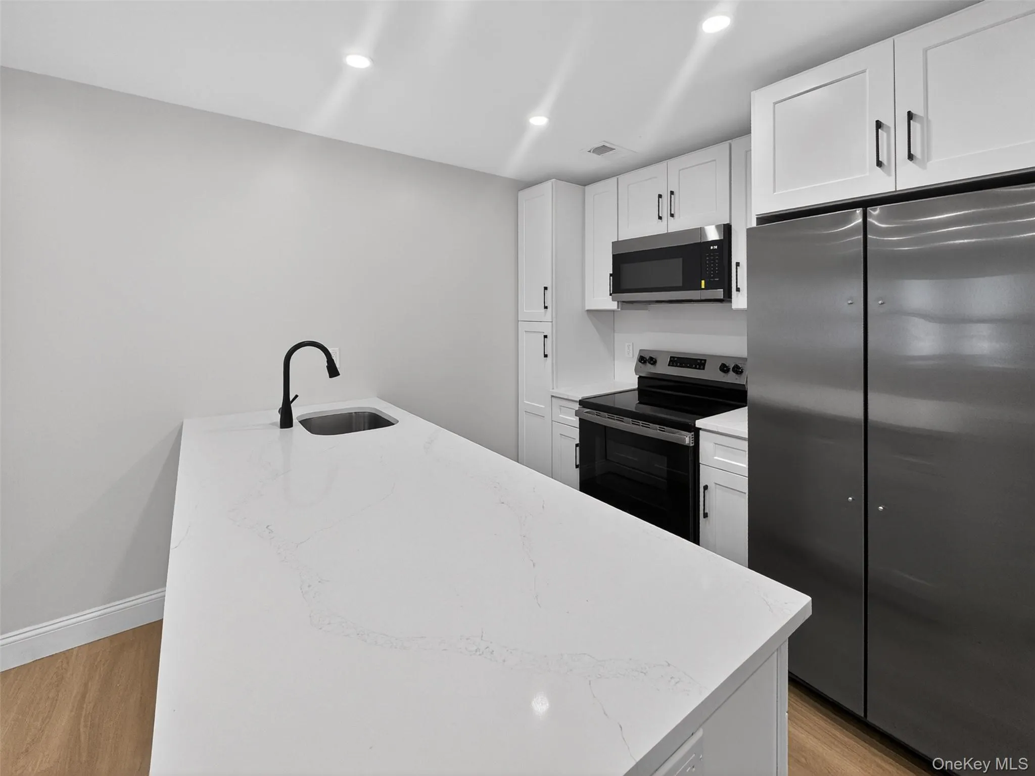 Kitchen featuring a large central island with an integrated sink and matte black faucet, white shaker-style cabinetry with dark hardware, stainless steel appliances, and recessed lighting Kitchen featuring a large central island with an integrated sink and matte black faucet, white shaker-style cabinetry with dark hardware, stainless steel appliances, and recessed lighting
