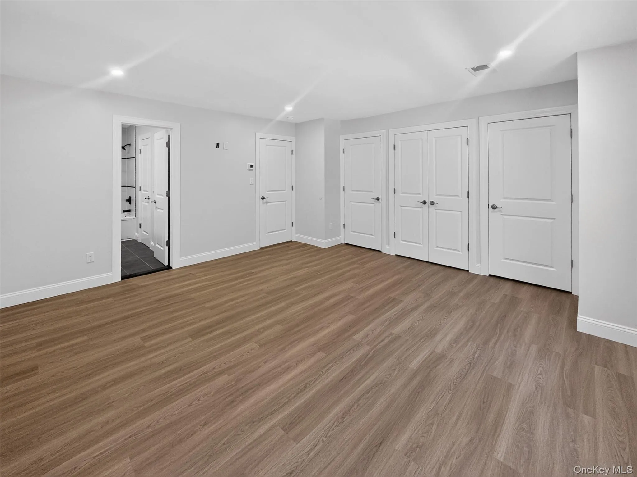Spacious room featuring wood-finish flooring, recessed lighting, multiple white panel doors, and a partial view of an adjacent bathroom with dark tile flooring Spacious room featuring wood-finish flooring, recessed lighting, multiple white panel doors, and a partial view of an adjacent bathroom with dark tile flooring