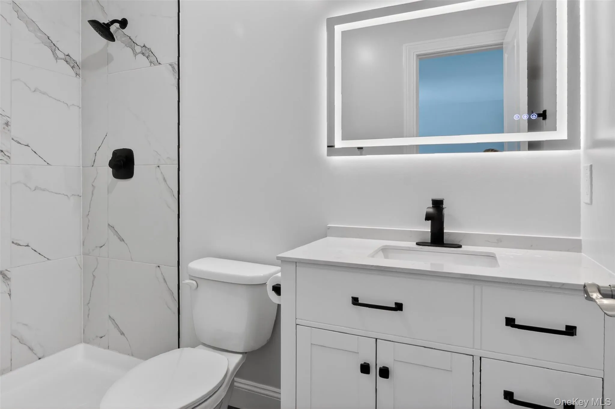Lower level - Bathroom featuring a lighted mirror, single-basin vanity with white cabinetry, matte black fixtures, and a shower with marble-look tile surround Lower level - Bathroom featuring a lighted mirror, single-basin vanity with white cabinetry, matte black fixtures, and a shower with marble-look tile surround