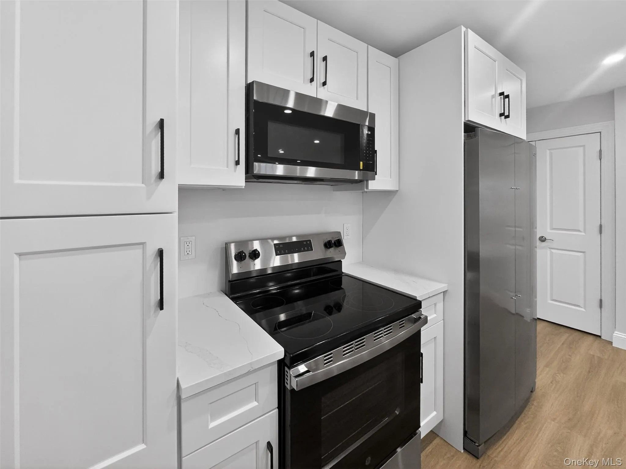 Kitchen featuring white shaker-style cabinetry, matte black hardware, stainless steel appliances, white countertops, and wood-finish flooring Kitchen featuring white shaker-style cabinetry, matte black hardware, stainless steel appliances, white countertops, and wood-finish flooring
