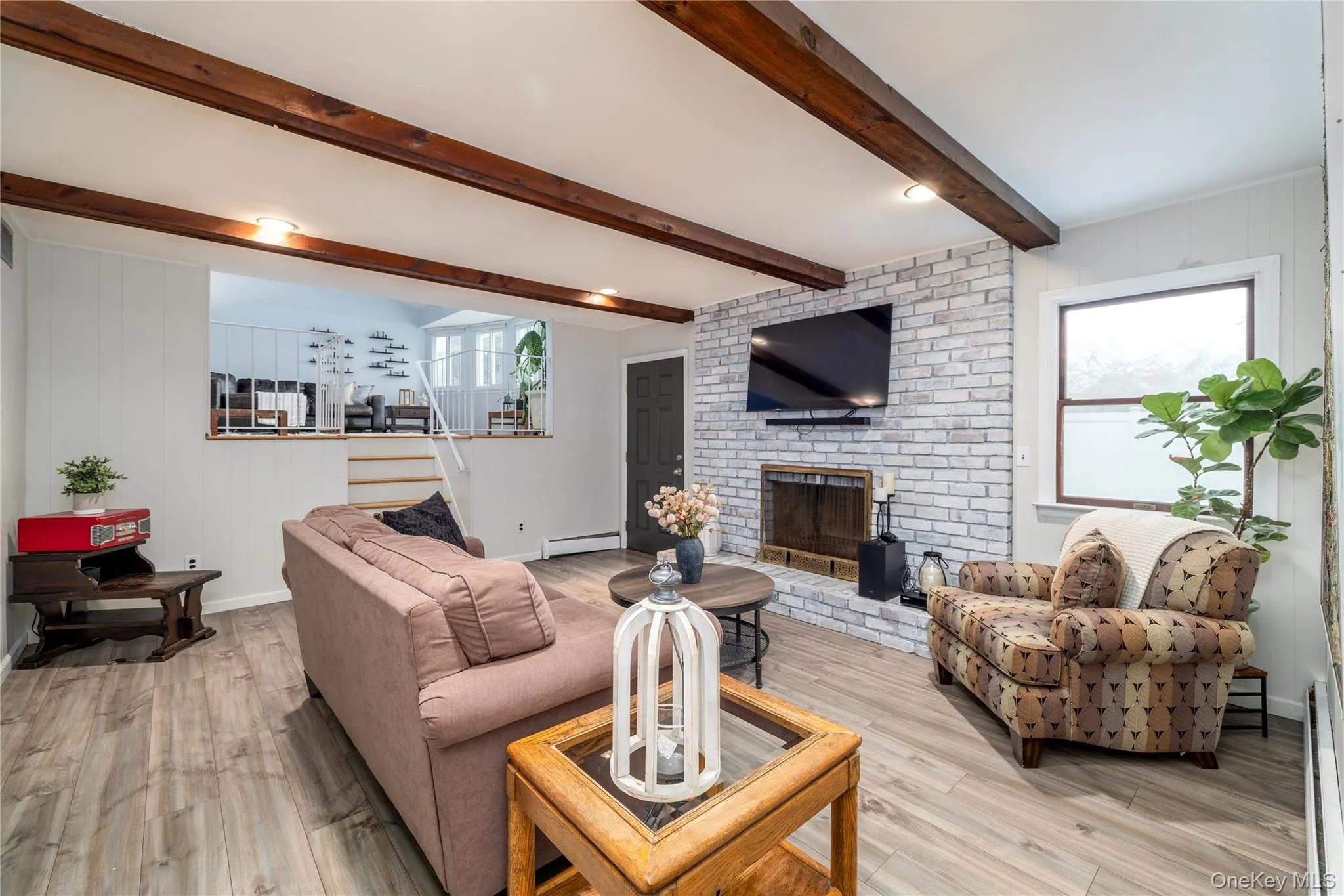 Living room featuring baseboards, a brick fireplace, light wood-type flooring, baseboard heating, and beamed ceiling Living room featuring baseboards, a brick fireplace, light wood-type flooring, baseboard heating, and beamed ceiling