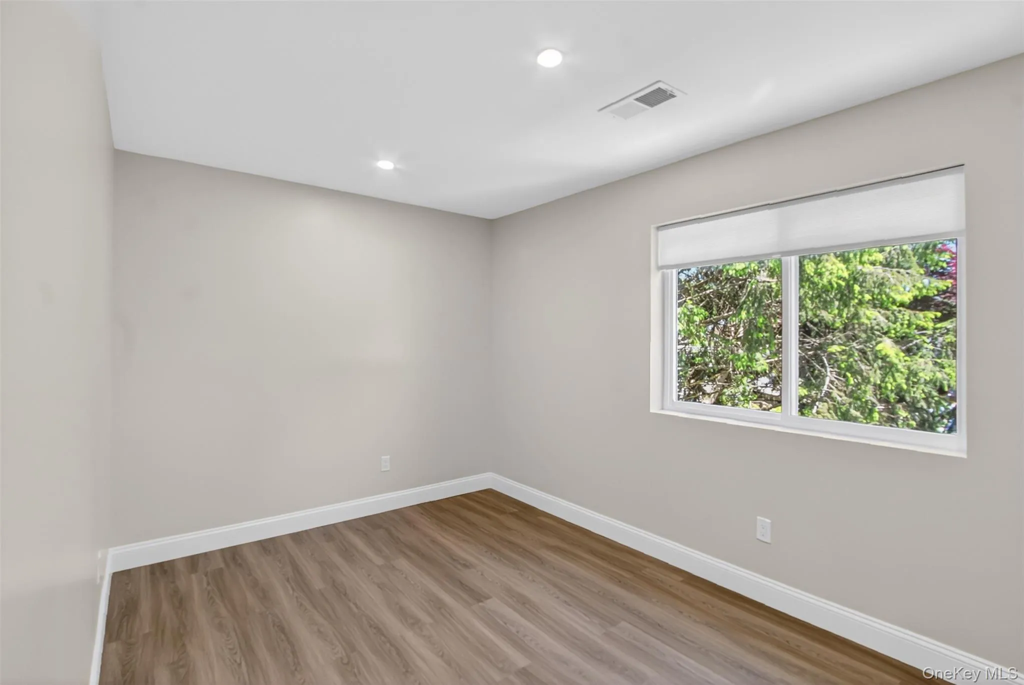 Room featuring a large window with roller shades, wood-finish flooring, recessed lighting, and light-toned walls with white baseboards Room featuring a large window with roller shades, wood-finish flooring, recessed lighting, and light-toned walls with white baseboards