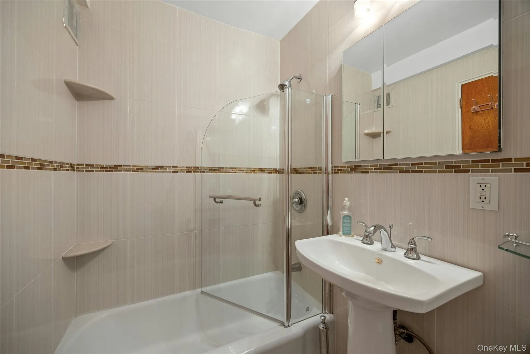 Bathroom featuring full wall tiling with a decorative mosaic band, a pedestal sink with chrome fixtures, and a bathtub with a partial glass enclosure Bathroom featuring full wall tiling with a decorative mosaic band, a pedestal sink with chrome fixtures, and a bathtub with a partial glass enclosure