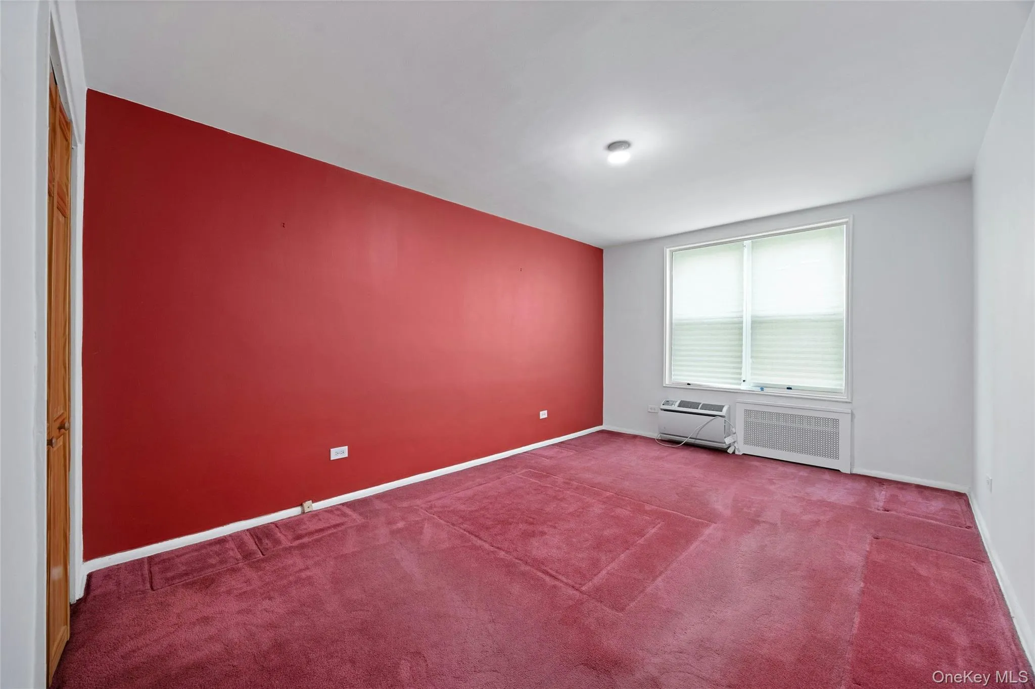 Spacious room featuring a large window with horizontal blinds, built-in air conditioning unit, and a red accent wall Spacious room featuring a large window with horizontal blinds, built-in air conditioning unit, and a red accent wall
