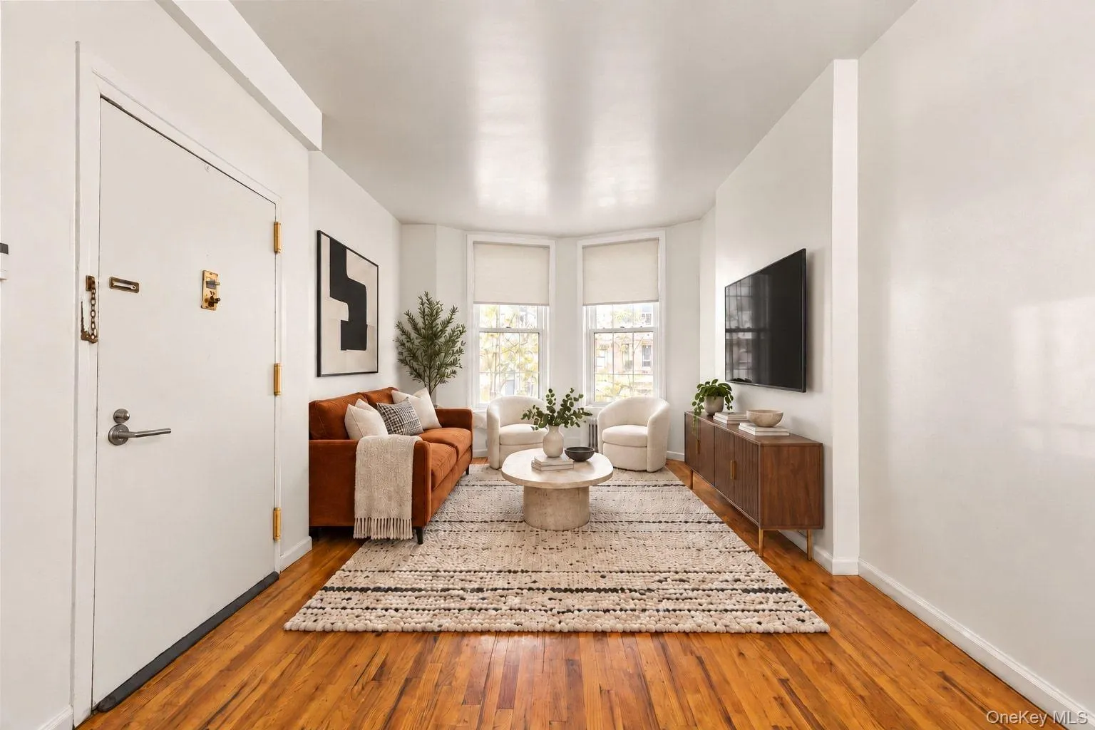 Hardwood flooring, bay window with roller shades, recessed wall space for media, and a built-in wood-finish console Hardwood flooring, bay window with roller shades, recessed wall space for media, and a built-in wood-finish console