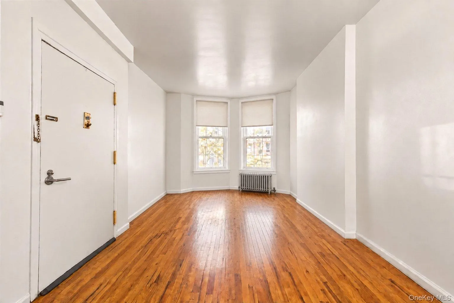 Hardwood flooring, painted white walls, white trim, two windows with roller shades, and a baseboard radiator Hardwood flooring, painted white walls, white trim, two windows with roller shades, and a baseboard radiator
