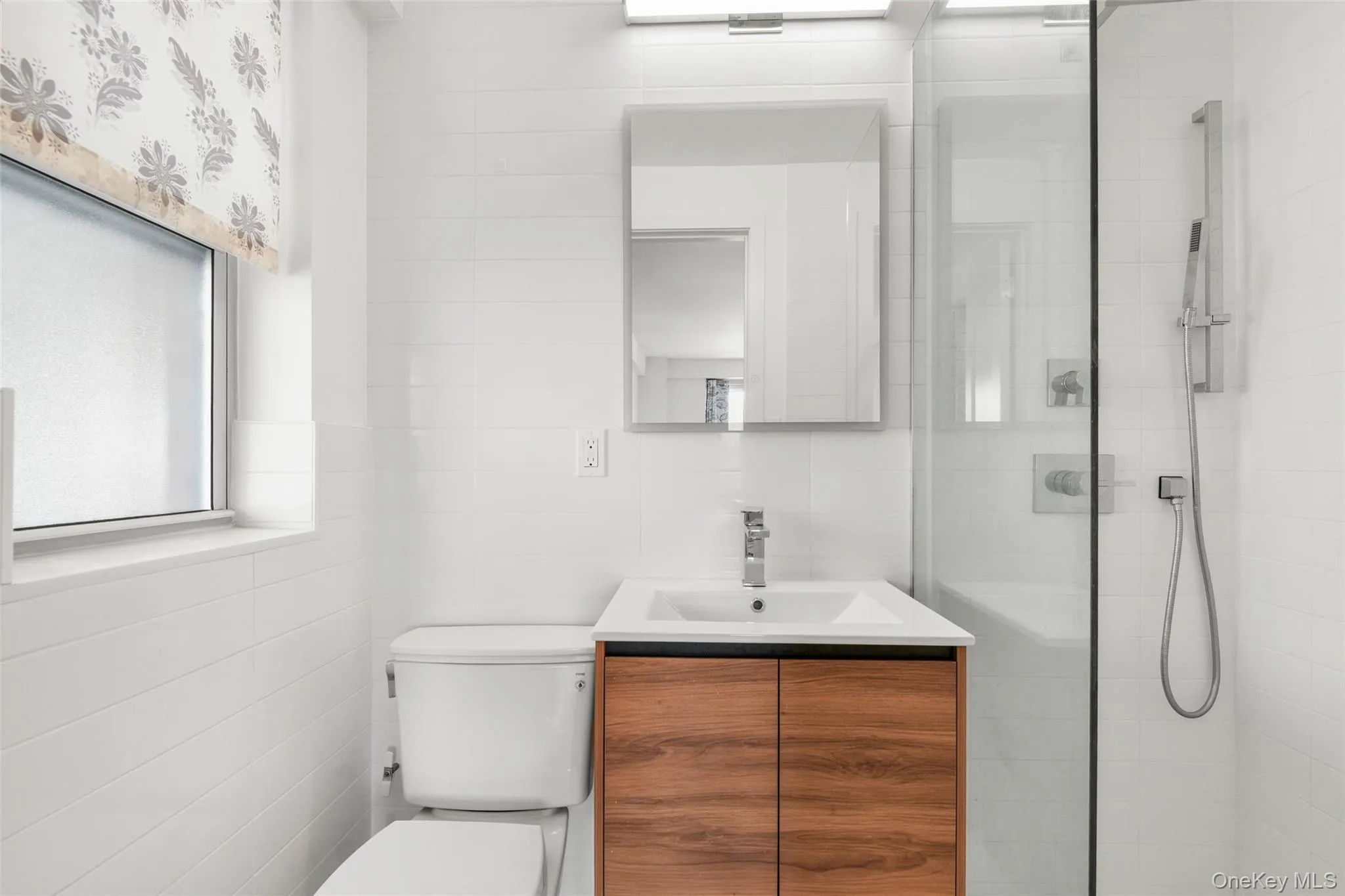 Tiled bathroom with a wood-finish vanity, integrated sink, mirrored medicine cabinet, and glass-enclosed shower Tiled bathroom with a wood-finish vanity, integrated sink, mirrored medicine cabinet, and glass-enclosed shower