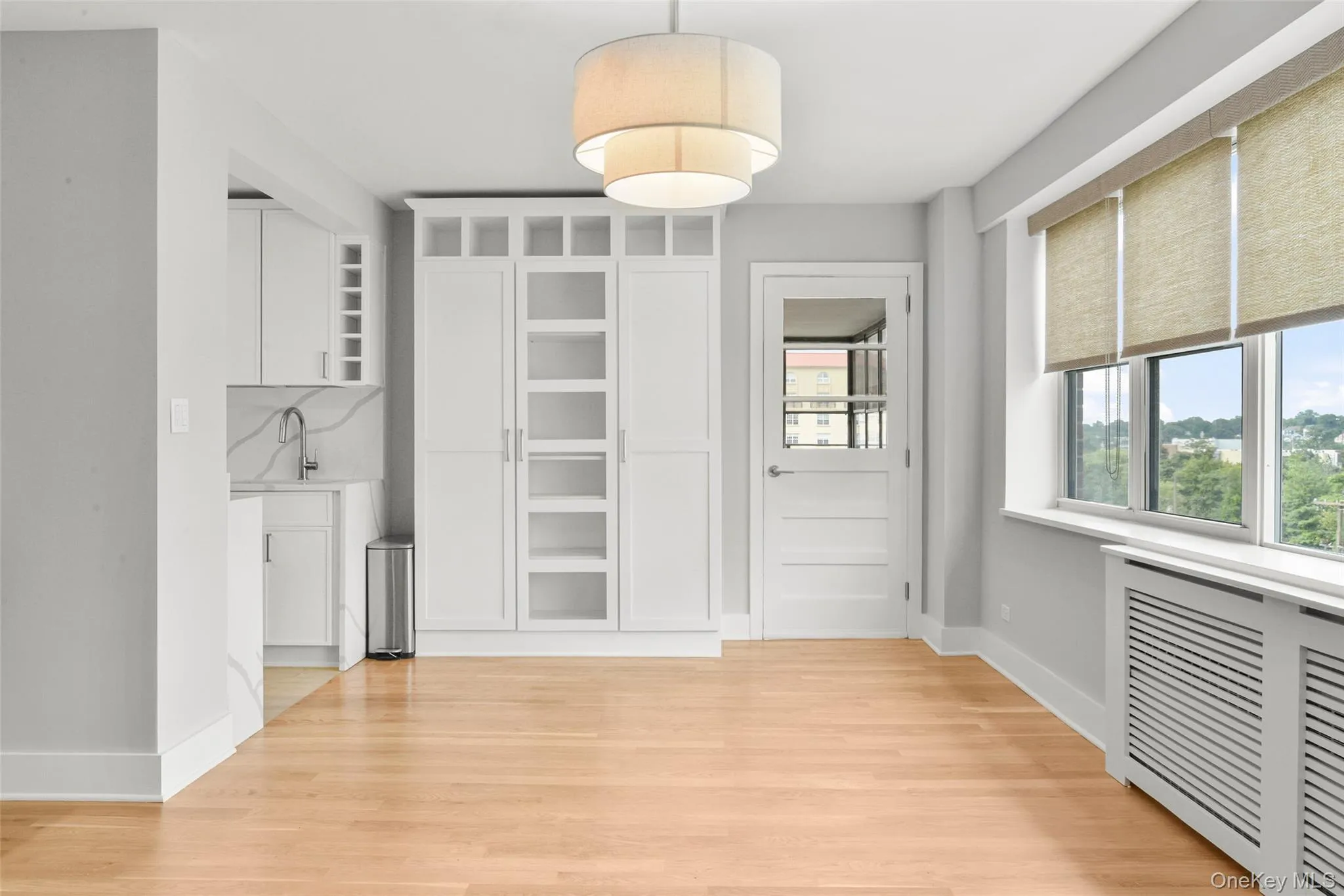 White built-in cabinetry with glass paneling, a contemporary tiered ceiling light fixture, and windows equipped with roller shades White built-in cabinetry with glass paneling, a contemporary tiered ceiling light fixture, and windows equipped with roller shades