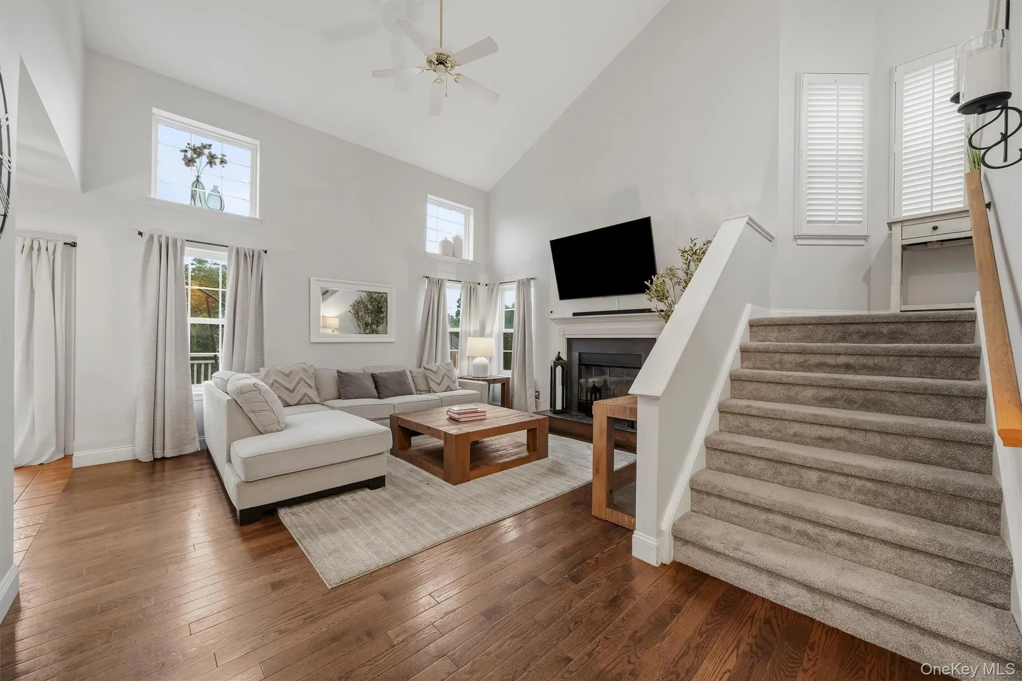 Hardwood-finish flooring, vaulted ceiling, fireplace with mantel, ceiling fan, and carpeted stairs with wood railing Hardwood-finish flooring, vaulted ceiling, fireplace with mantel, ceiling fan, and carpeted stairs with wood railing