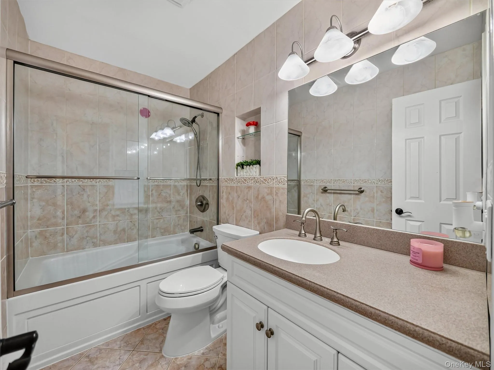 Bathtub with sliding glass enclosure, tile surround, brushed nickel fixtures, vanity with integrated sink, and large mirror with overhead light bar Bathtub with sliding glass enclosure, tile surround, brushed nickel fixtures, vanity with integrated sink, and large mirror with overhead light bar