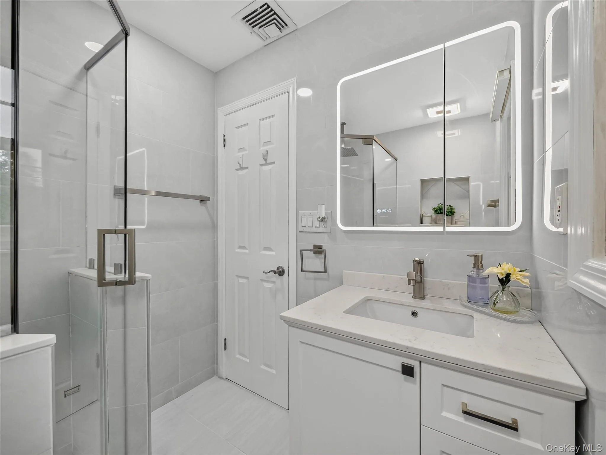 Glass-enclosed shower with a modern handle, white vanity with a rectangular sink, stone-finish countertop, and a backlit medicine cabinet Glass-enclosed shower with a modern handle, white vanity with a rectangular sink, stone-finish countertop, and a backlit medicine cabinet