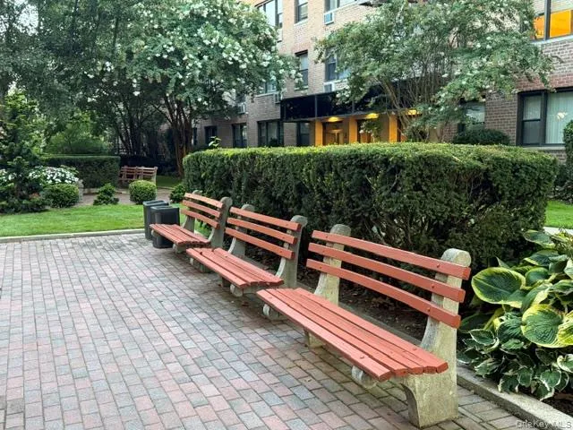 Paved outdoor area with three wooden and concrete benches, manicured hedges, and lush landscaping Paved outdoor area with three wooden and concrete benches, manicured hedges, and lush landscaping