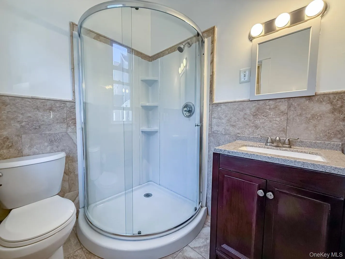 Curved glass shower enclosure with built-in shelving, vanity with dark wood-finish cabinetry, and a light-toned countertop Curved glass shower enclosure with built-in shelving, vanity with dark wood-finish cabinetry, and a light-toned countertop