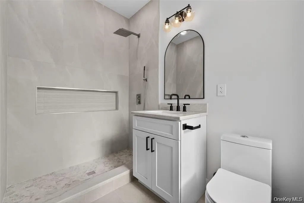 Walk-in shower with rain head and tiled niche, white vanity with stone countertop, black-framed arched mirror, and contemporary vanity light fixture Walk-in shower with rain head and tiled niche, white vanity with stone countertop, black-framed arched mirror, and contemporary vanity light fixture