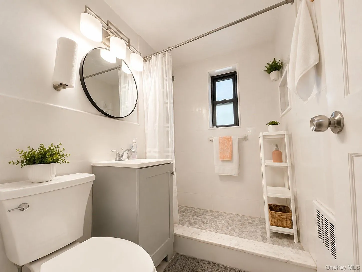 Bathroom featuring a gray vanity with a white countertop, round wall mirror, modern three-light fixture, and a shower with pebble tile flooring Bathroom featuring a gray vanity with a white countertop, round wall mirror, modern three-light fixture, and a shower with pebble tile flooring