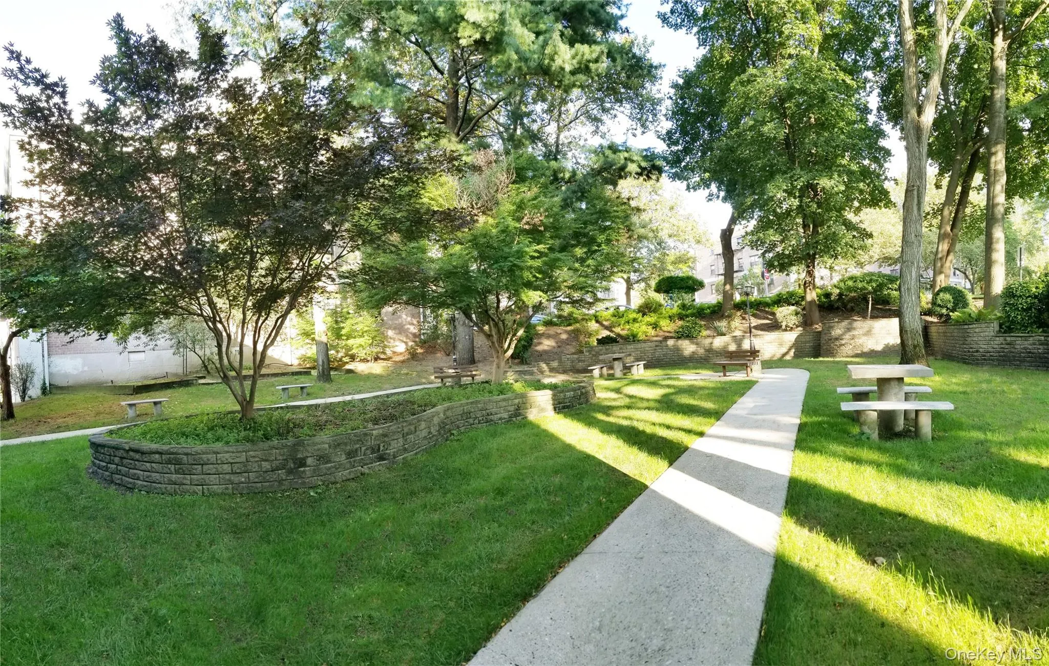 Expansive green space featuring mature trees, a concrete pathway, raised garden beds with stone retaining walls, and multiple stone picnic tables Expansive green space featuring mature trees, a concrete pathway, raised garden beds with stone retaining walls, and multiple stone picnic tables