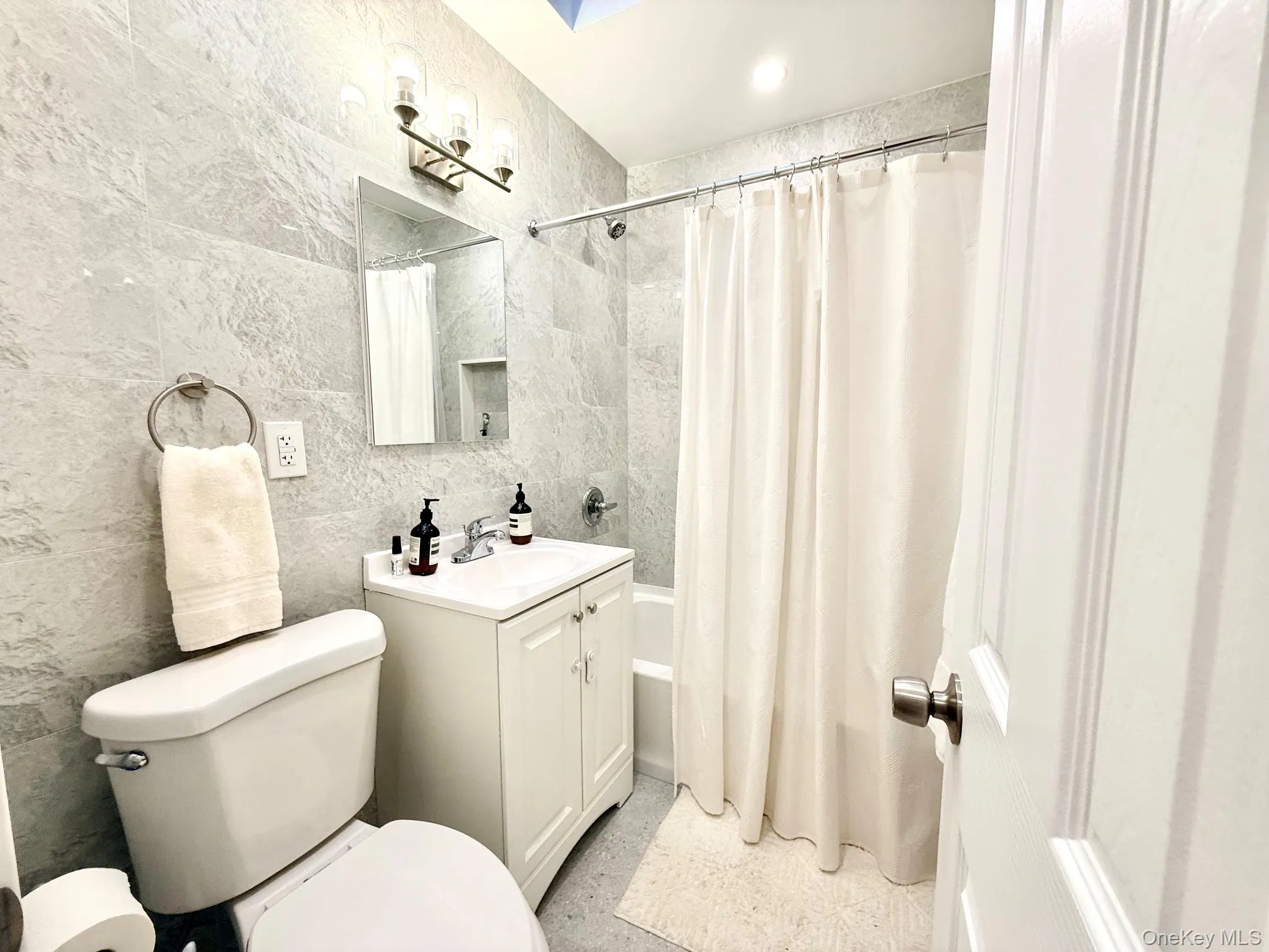 Modern bathroom featuring light-toned wall tiling, a white vanity with an integrated sink, a brushed nickel light fixture, a white toilet, and a white tub with shower Modern bathroom featuring light-toned wall tiling, a white vanity with an integrated sink, a brushed nickel light fixture, a white toilet, and a white tub with shower