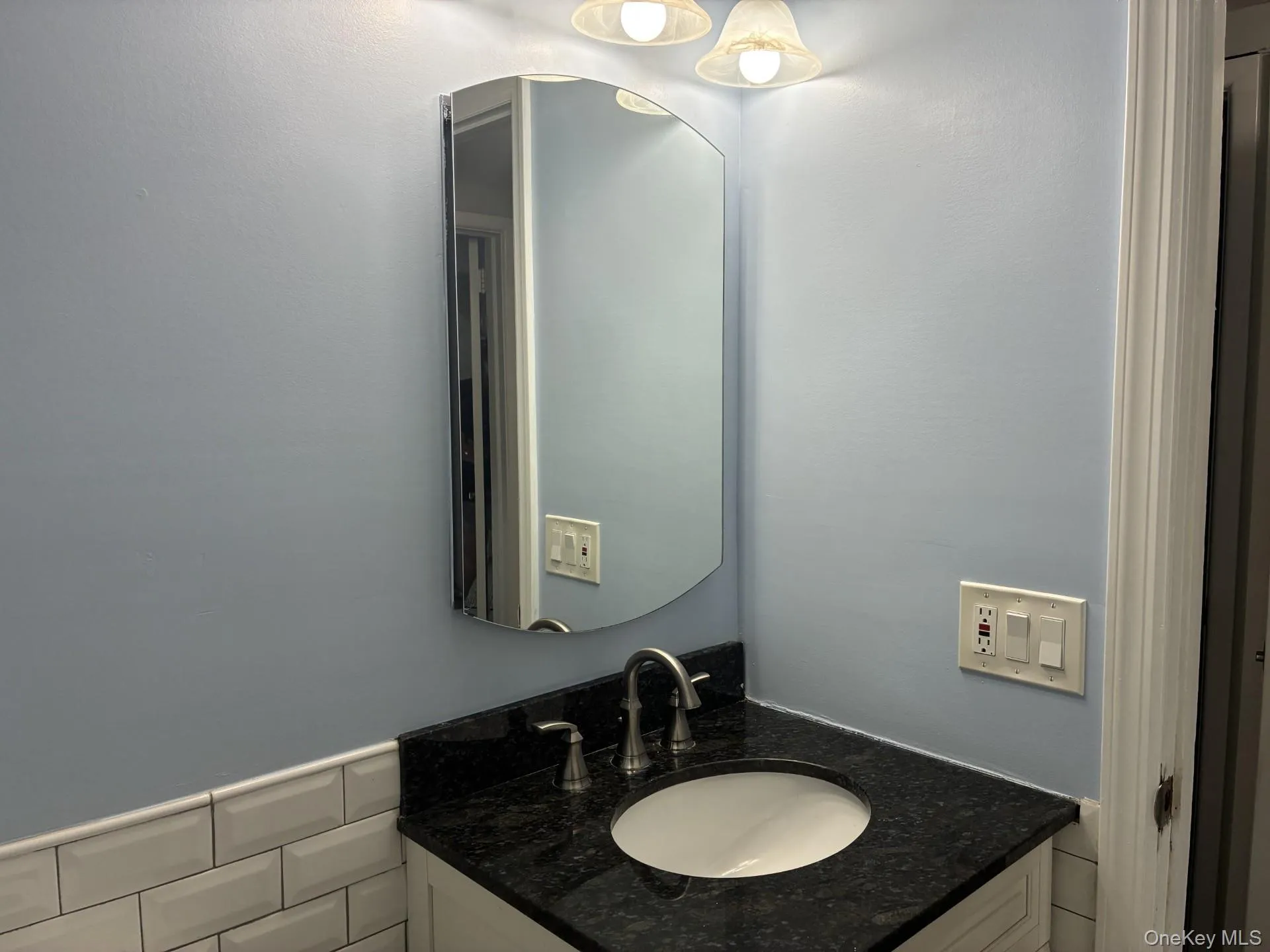 Vanity with dark countertop and undermount sink, wall-mounted light fixture, and subway tile wainscoting Vanity with dark countertop and undermount sink, wall-mounted light fixture, and subway tile wainscoting