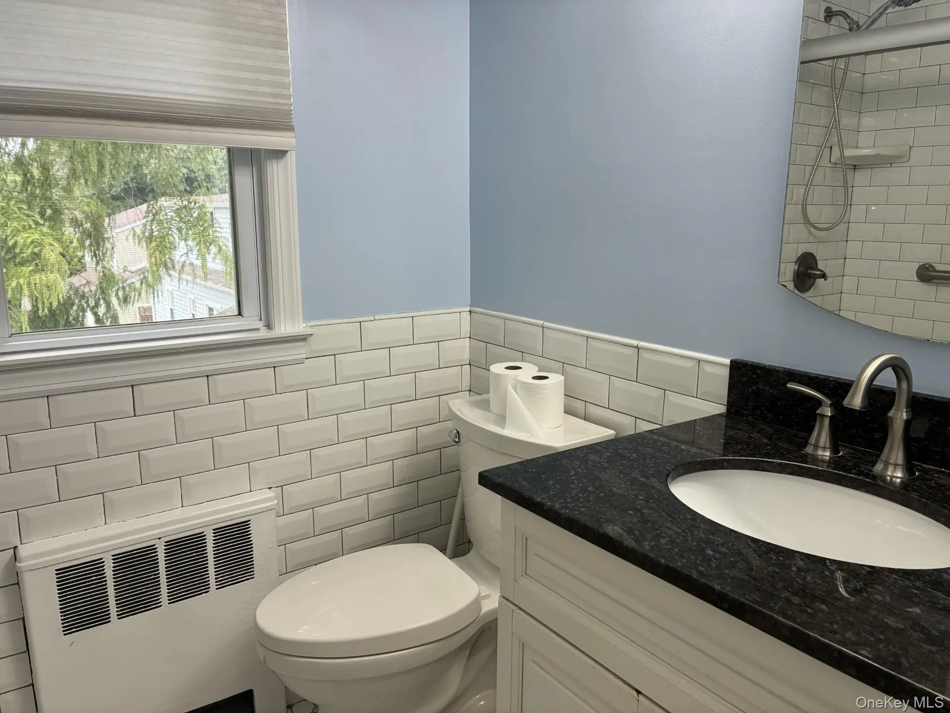 White subway tile wainscoting, solid surface countertop with integrated sink, modern vanity, window with natural lighting, and a wall-mounted mirror White subway tile wainscoting, solid surface countertop with integrated sink, modern vanity, window with natural lighting, and a wall-mounted mirror