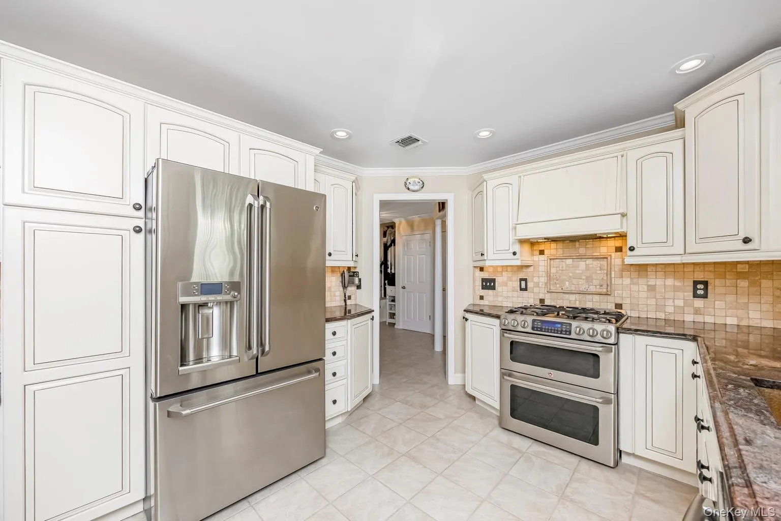 White shaker-style cabinetry, stainless steel French door refrigerator, stainless steel double oven range, mosaic tile backsplash, and light-toned tile flooring White shaker-style cabinetry, stainless steel French door refrigerator, stainless steel double oven range, mosaic tile backsplash, and light-toned tile flooring