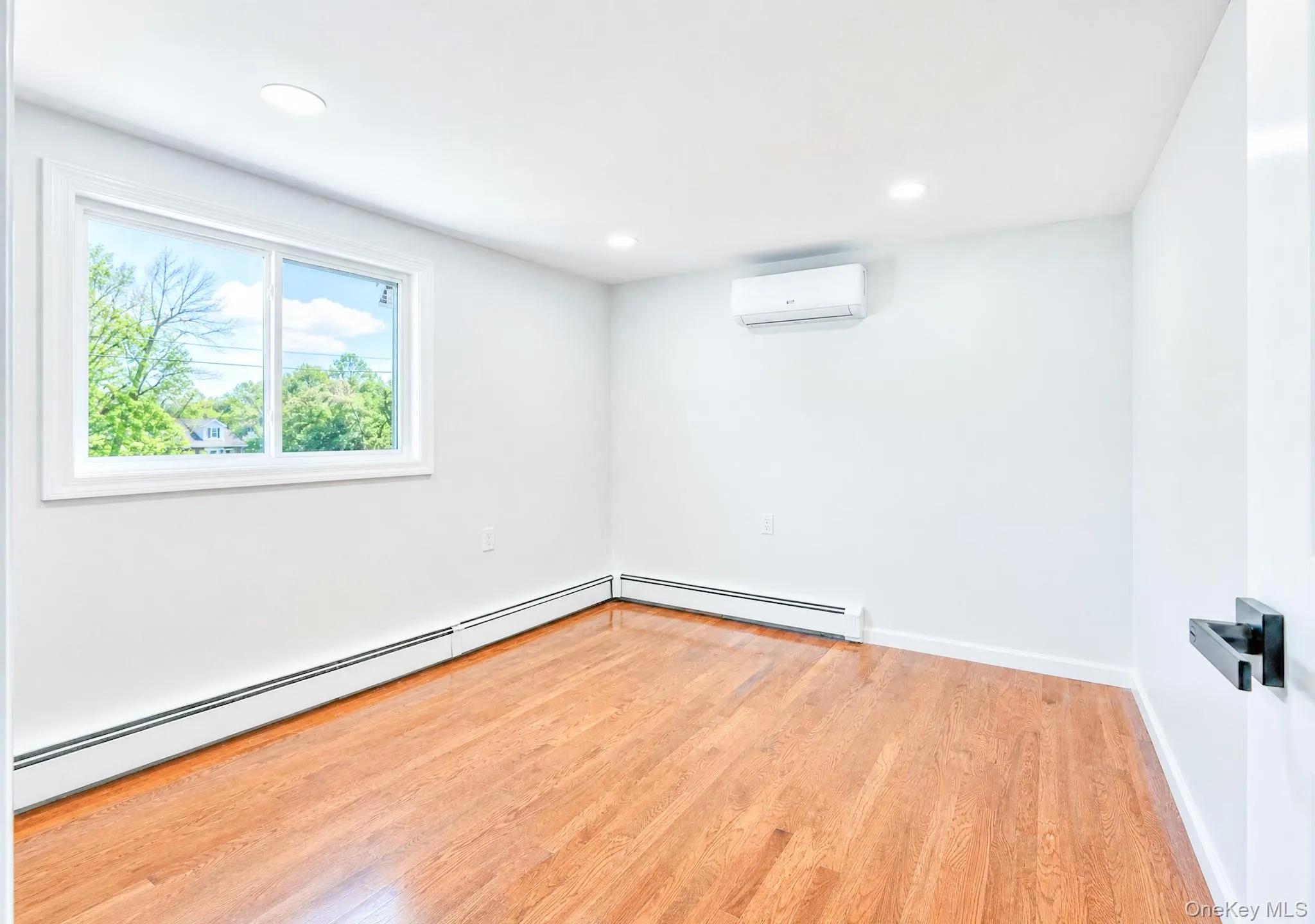 Light-filled room featuring wood-finish flooring, a white trim window, baseboard heating, recessed lighting, and a wall-mounted climate control unit Light-filled room featuring wood-finish flooring, a white trim window, baseboard heating, recessed lighting, and a wall-mounted climate control unit