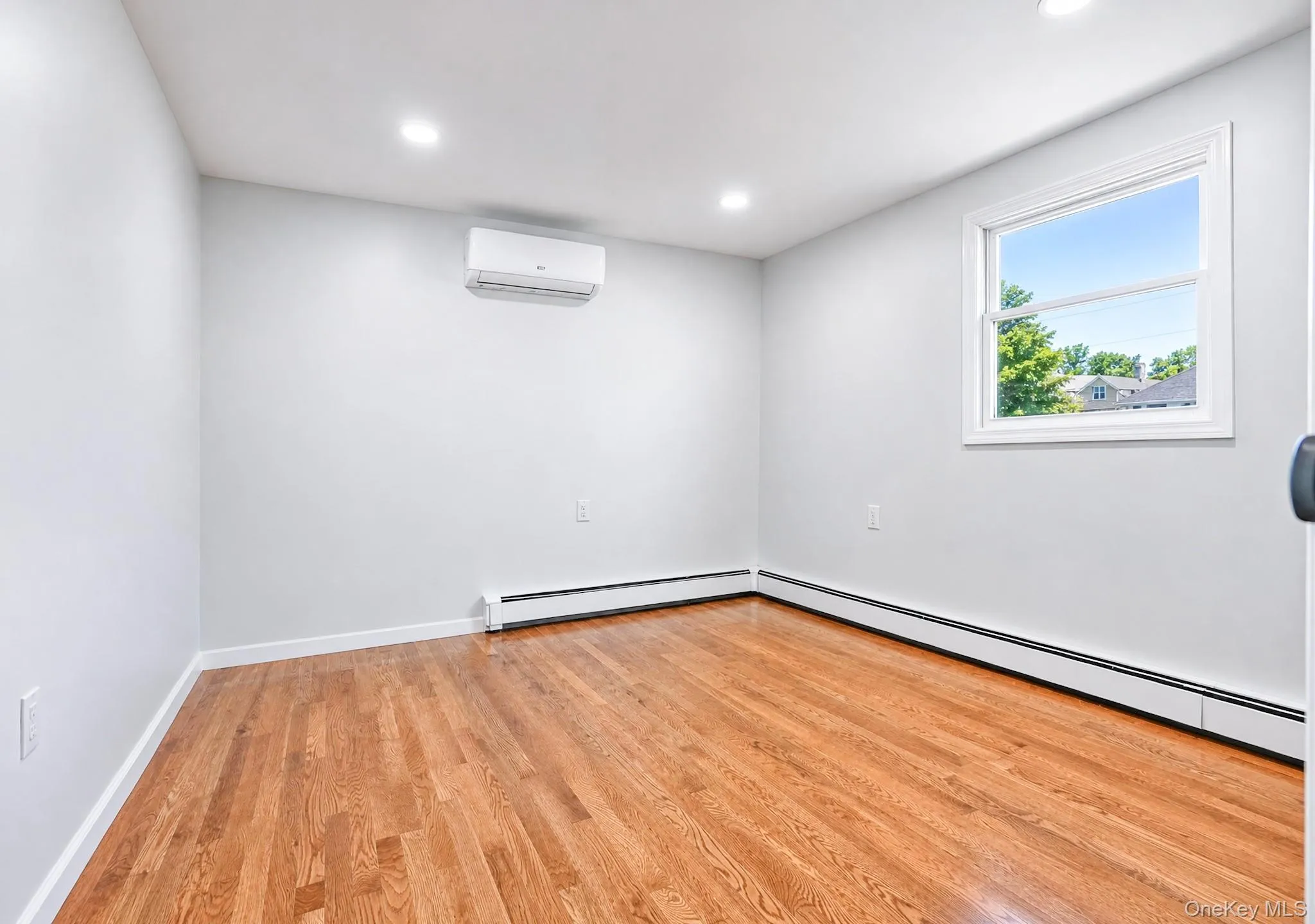 Room featuring hardwood flooring, white baseboards, recessed lighting, and a white-framed window Room featuring hardwood flooring, white baseboards, recessed lighting, and a white-framed window