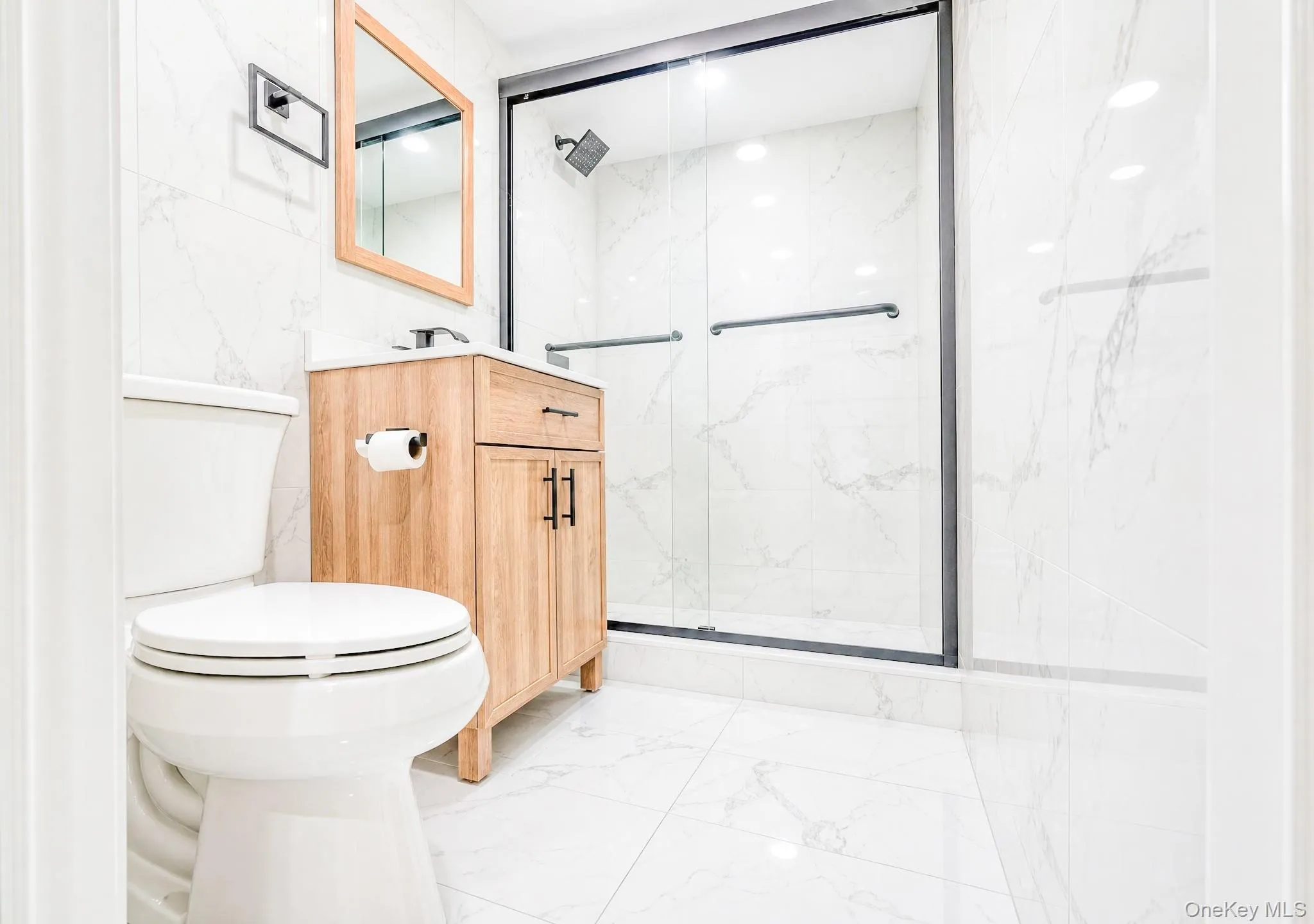 Bathroom featuring a frameless glass shower enclosure with matte black trim, a wood-finish vanity with ample storage, a rectangular wood-framed mirror, and white tile flooring and walls Bathroom featuring a frameless glass shower enclosure with matte black trim, a wood-finish vanity with ample storage, a rectangular wood-framed mirror, and white tile flooring and walls