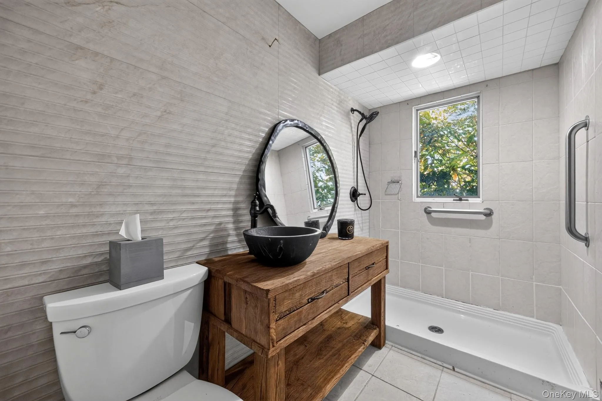 Textured tile wall, wood vanity with basin sink, walk-in shower with grab bars, and exterior window Textured tile wall, wood vanity with basin sink, walk-in shower with grab bars, and exterior window