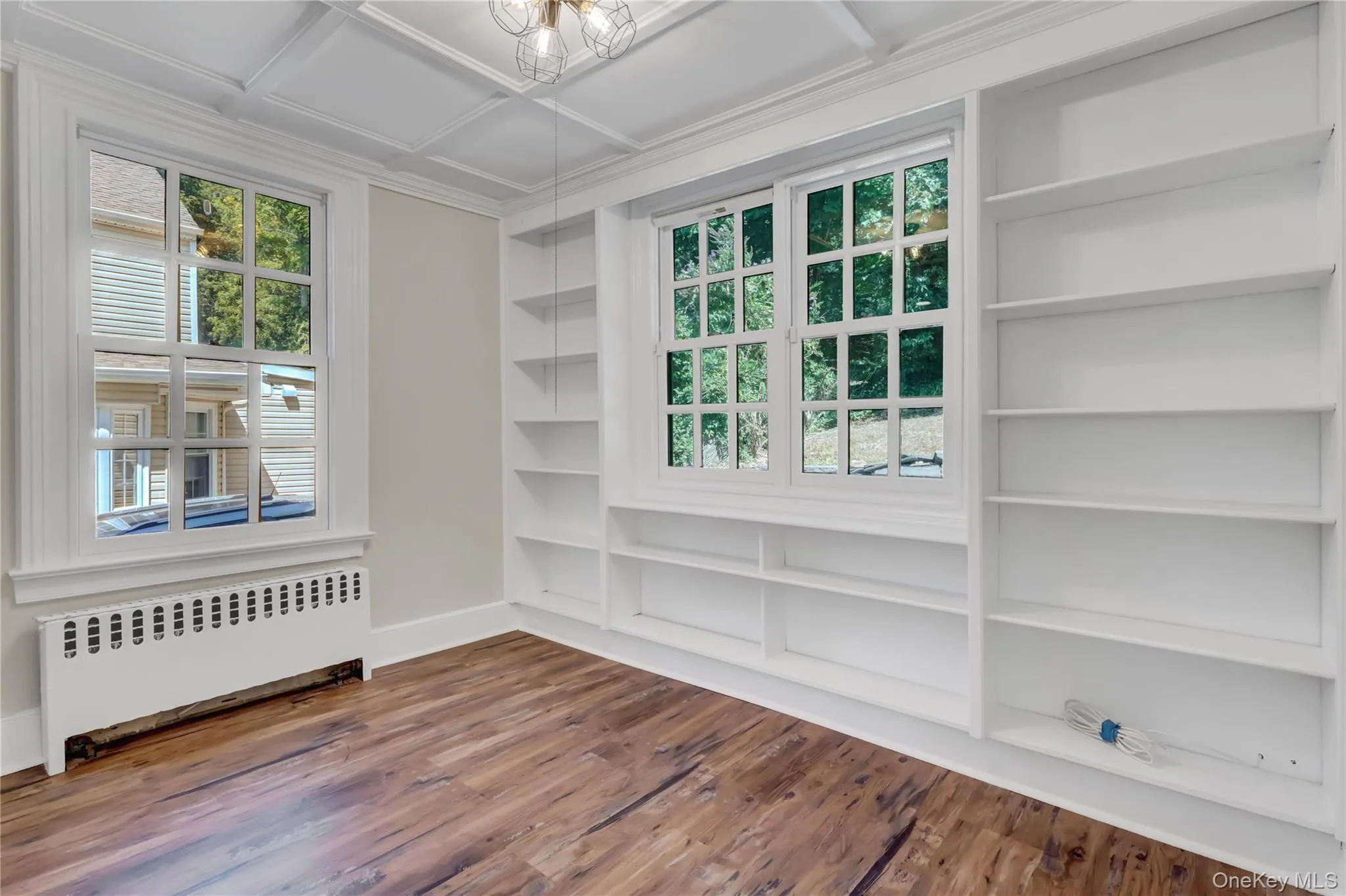 Empty room featuring radiator heating unit, built in shelves, coffered ceiling, wood finished floors, and a chandelier Empty room featuring radiator heating unit, built in shelves, coffered ceiling, wood finished floors, and a chandelier