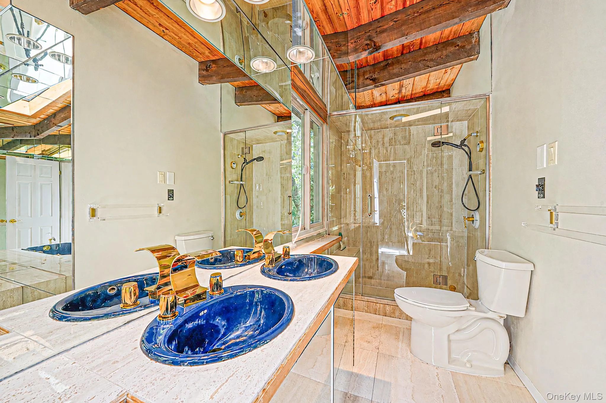 Dual vanity with blue vessel sinks and gold-tone fixtures, glass-enclosed shower, exposed wood beam ceiling, and skylight Dual vanity with blue vessel sinks and gold-tone fixtures, glass-enclosed shower, exposed wood beam ceiling, and skylight