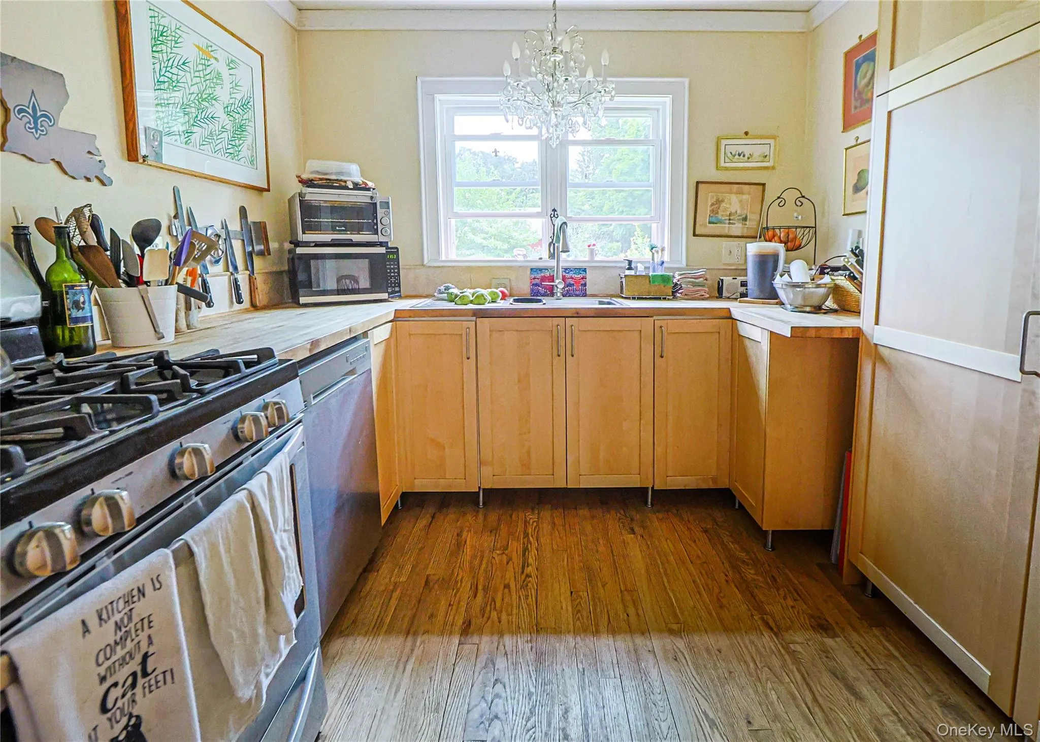 Kitchen featuring wood-finish flooring, light wood cabinetry, butcher block countertops, a stainless steel gas range, and a window with a chandelier Kitchen featuring wood-finish flooring, light wood cabinetry, butcher block countertops, a stainless steel gas range, and a window with a chandelier