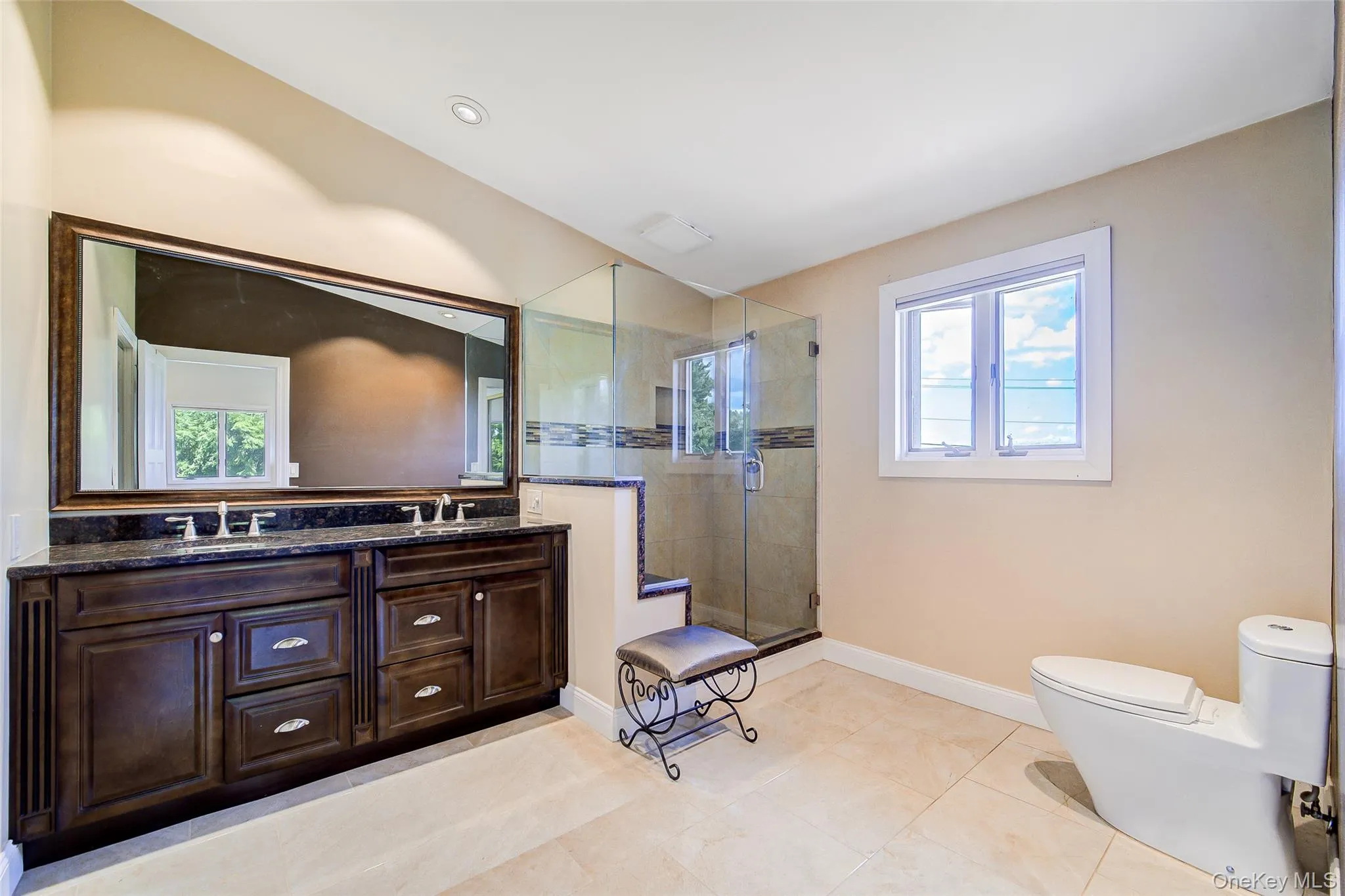 Dual vanity with dark wood cabinetry and stone countertop, large wall-mounted mirror, enclosed glass shower, recessed lighting, and a white framed window Dual vanity with dark wood cabinetry and stone countertop, large wall-mounted mirror, enclosed glass shower, recessed lighting, and a white framed window
