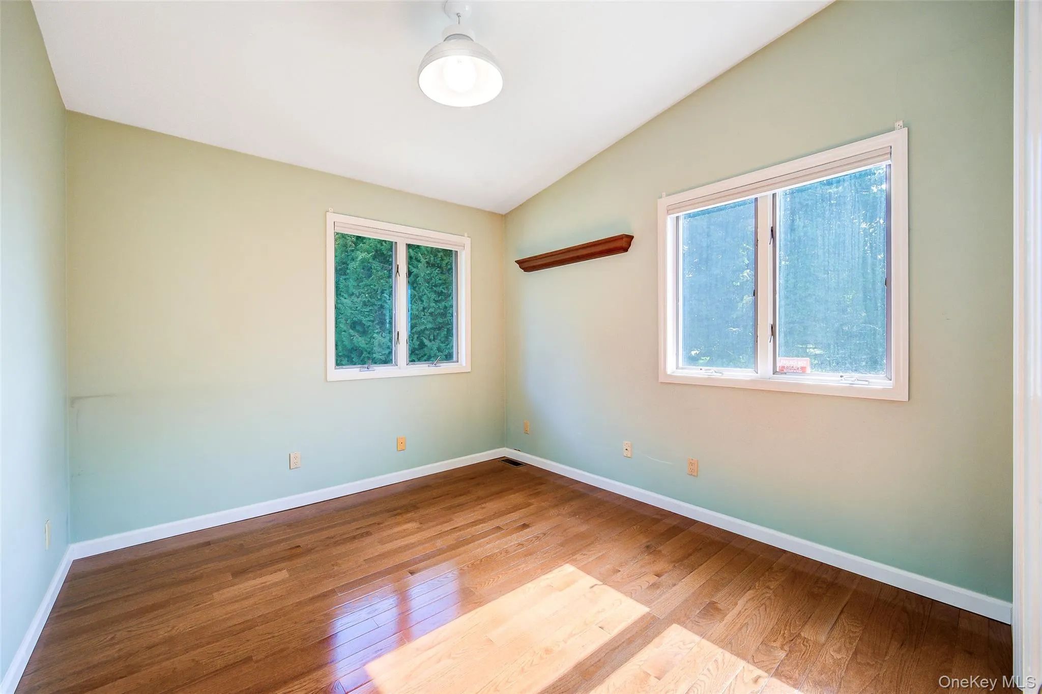 Vaulted ceiling with a flush-mount light fixture, two windows, wood-finish flooring, and a wall-mounted wooden shelf Vaulted ceiling with a flush-mount light fixture, two windows, wood-finish flooring, and a wall-mounted wooden shelf