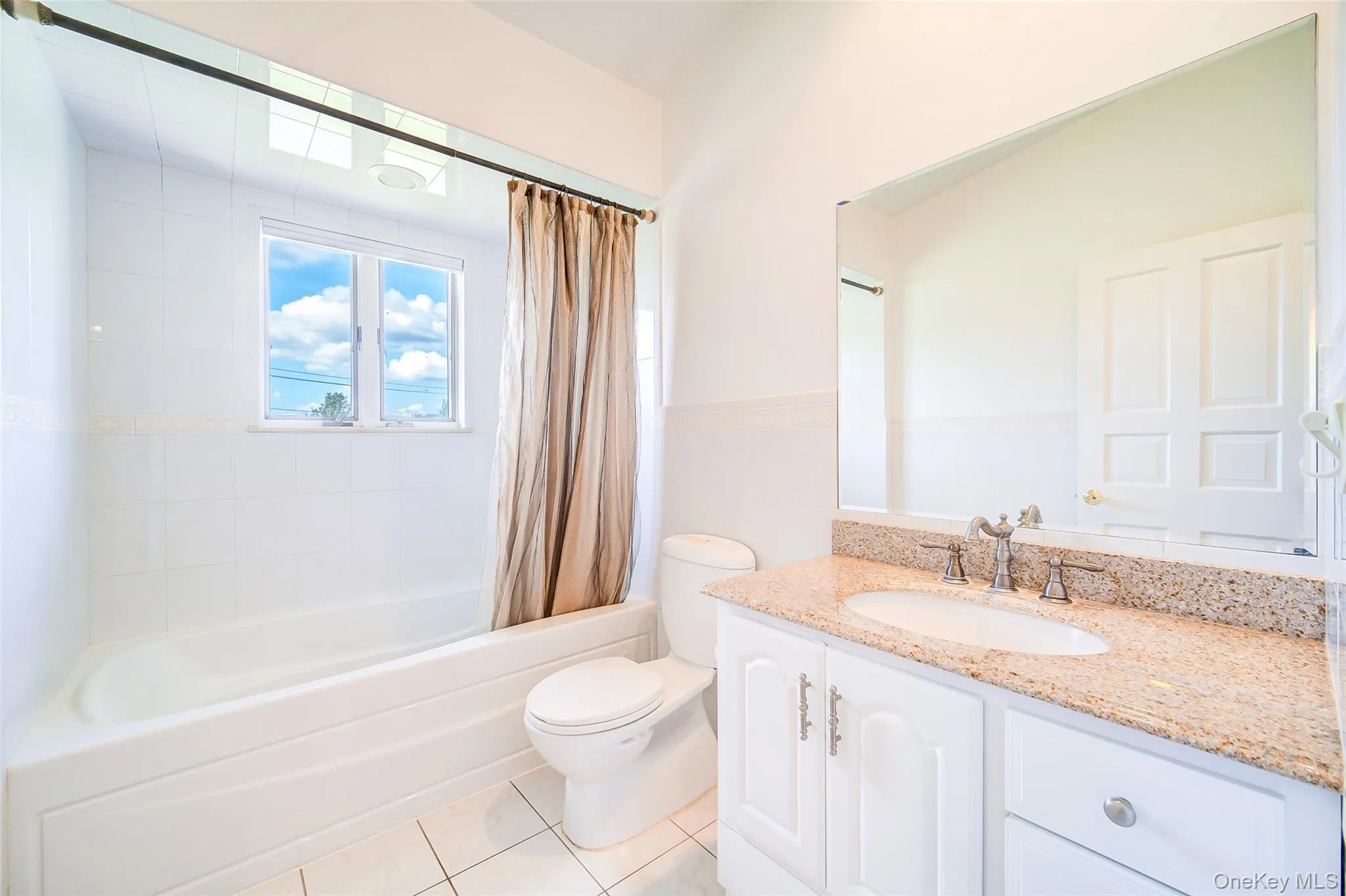 Full bath has tile flooring, white vanity with stone countertop, undermount sink with dual-handle faucet, white shower/tub with tile surround, and an expansive wall mirror Full bath has tile flooring, white vanity with stone countertop, undermount sink with dual-handle faucet, white shower/tub with tile surround, and an expansive wall mirror