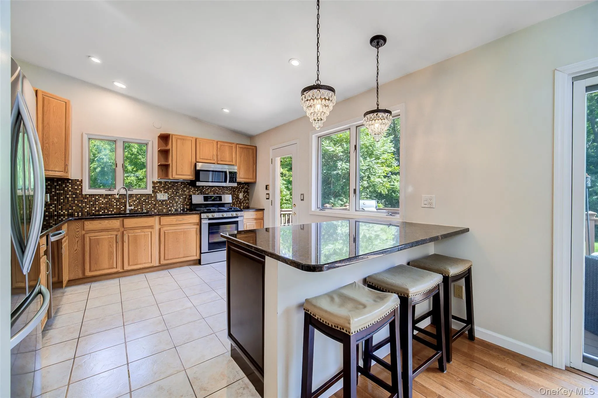 Kitchen featuring stainless steel appliances, wood-finish cabinetry, tile flooring, a dark countertop island with pendant lighting, and multiple windows Kitchen featuring stainless steel appliances, wood-finish cabinetry, tile flooring, a dark countertop island with pendant lighting, and multiple windows