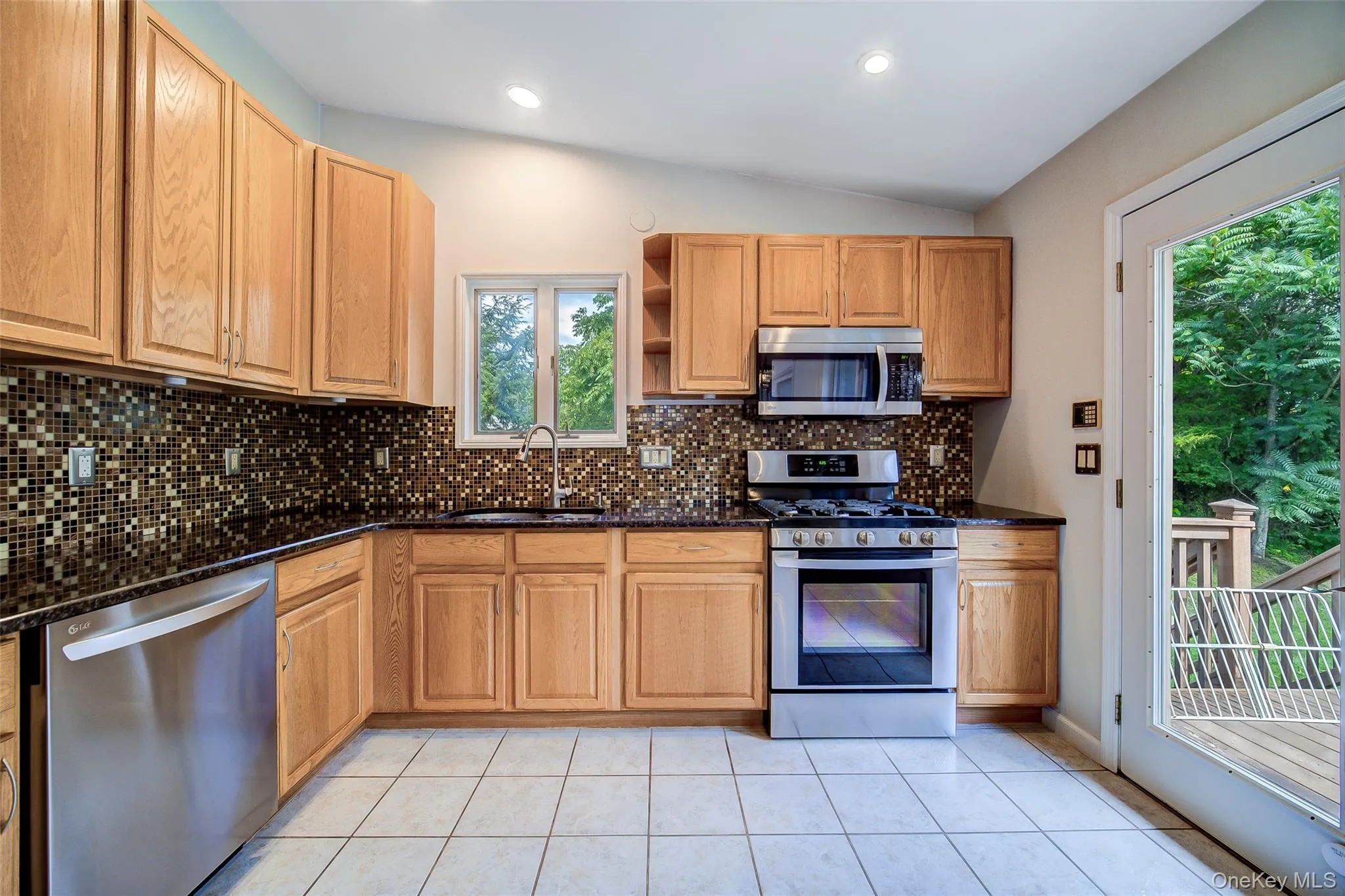Wood-finish cabinetry, stainless steel appliances, mosaic tile backsplash, black countertops, and white tiled flooring Wood-finish cabinetry, stainless steel appliances, mosaic tile backsplash, black countertops, and white tiled flooring