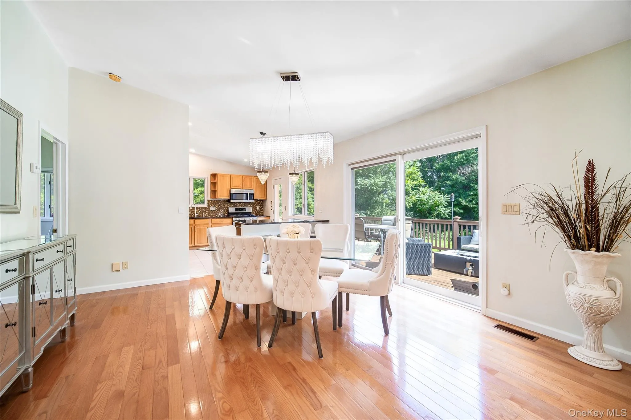 Hardwood flooring throughout, vaulted ceilings, modern linear chandelier, glass sliding door to an outdoor deck, and a kitchen visible with wood cabinetry Hardwood flooring throughout, vaulted ceilings, modern linear chandelier, glass sliding door to an outdoor deck, and a kitchen visible with wood cabinetry