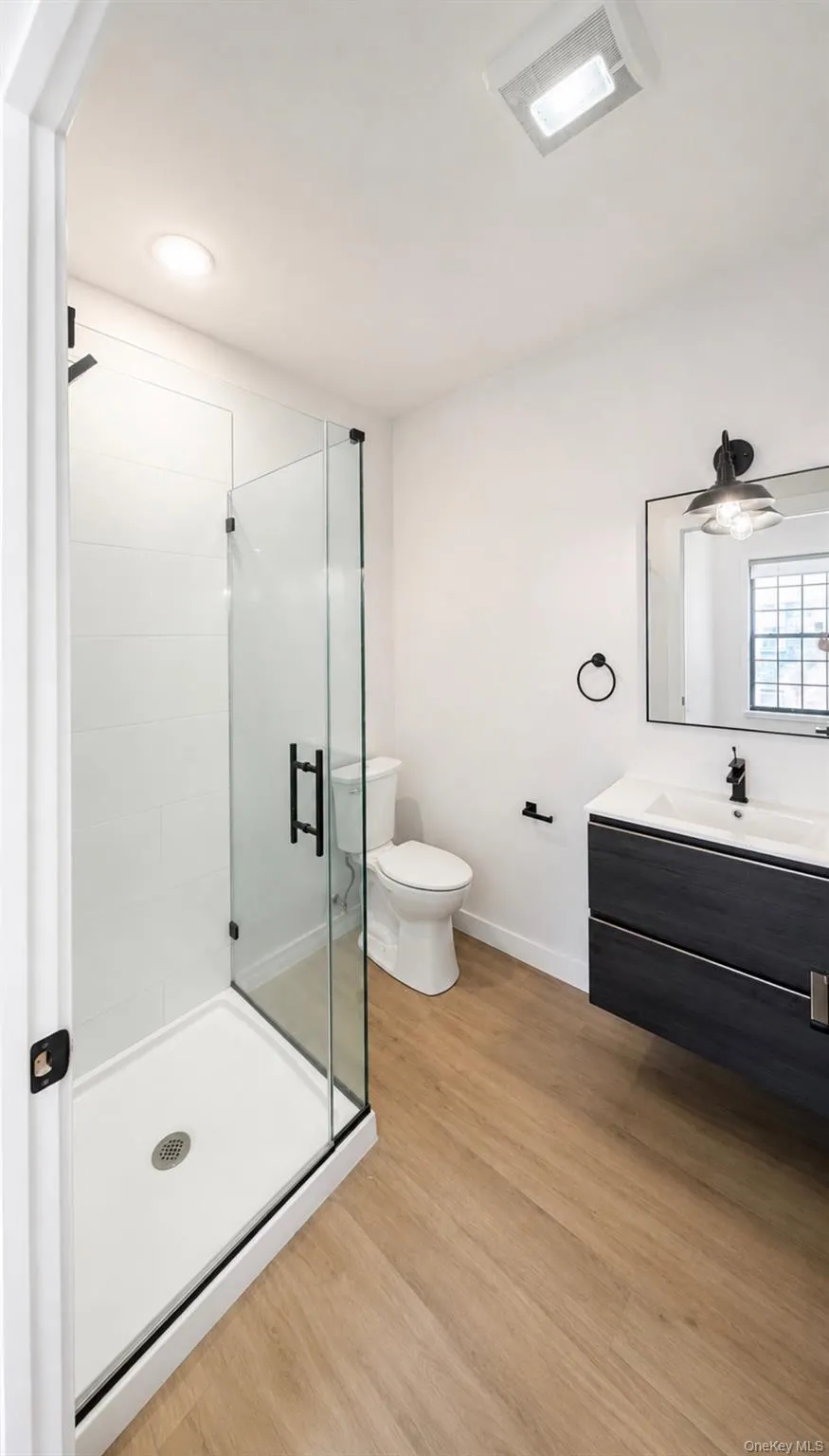 Walk-in shower with clear glass enclosure, floating vanity with integrated sink, wood-finish flooring, and industrial-style lighting fixture Walk-in shower with clear glass enclosure, floating vanity with integrated sink, wood-finish flooring, and industrial-style lighting fixture