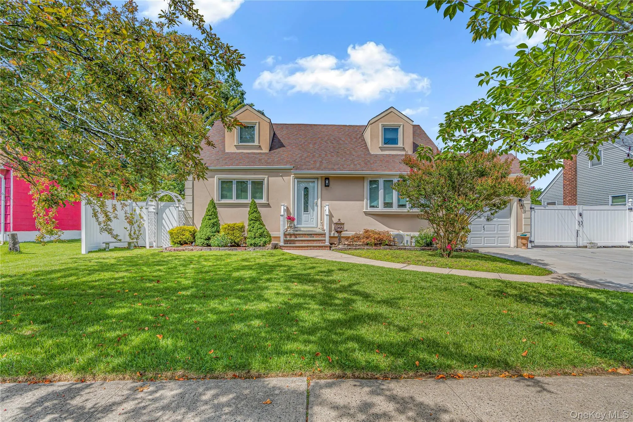 Stucco exterior with dormer windows, a concrete driveway, and a white fence Stucco exterior with dormer windows, a concrete driveway, and a white fence